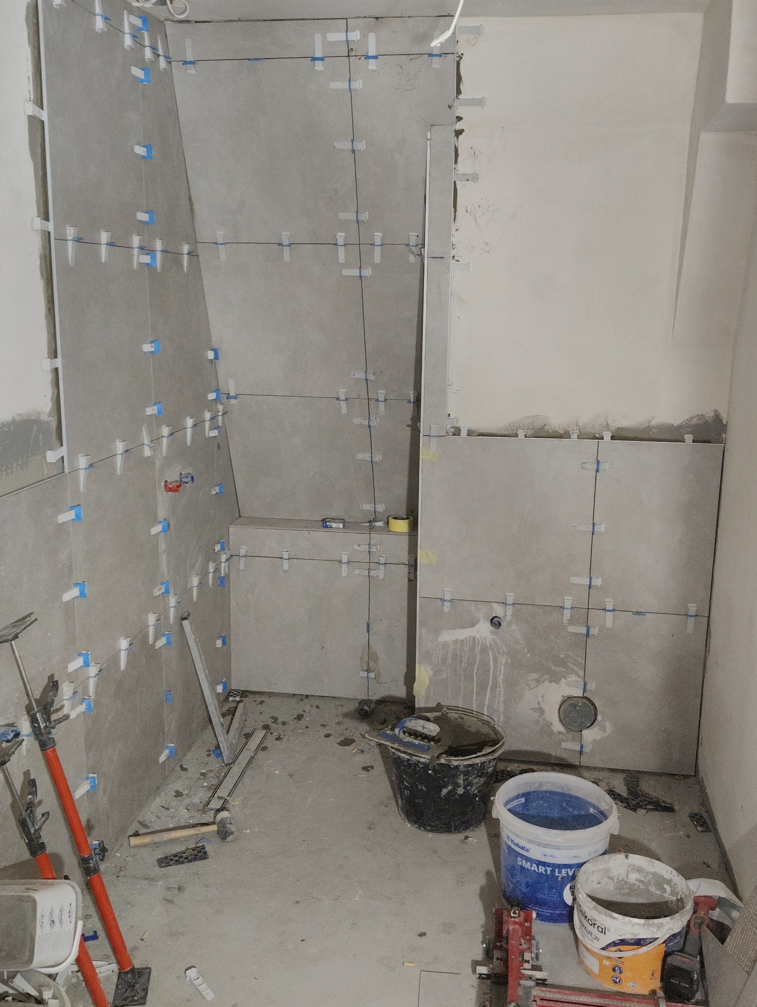 Szare płytki ścienne w trakcie montażu w łazience. Widoczne narzędzia glazurnicze, system poziomowania płytek i fragmenty ścian przygotowane do dalszych prac.