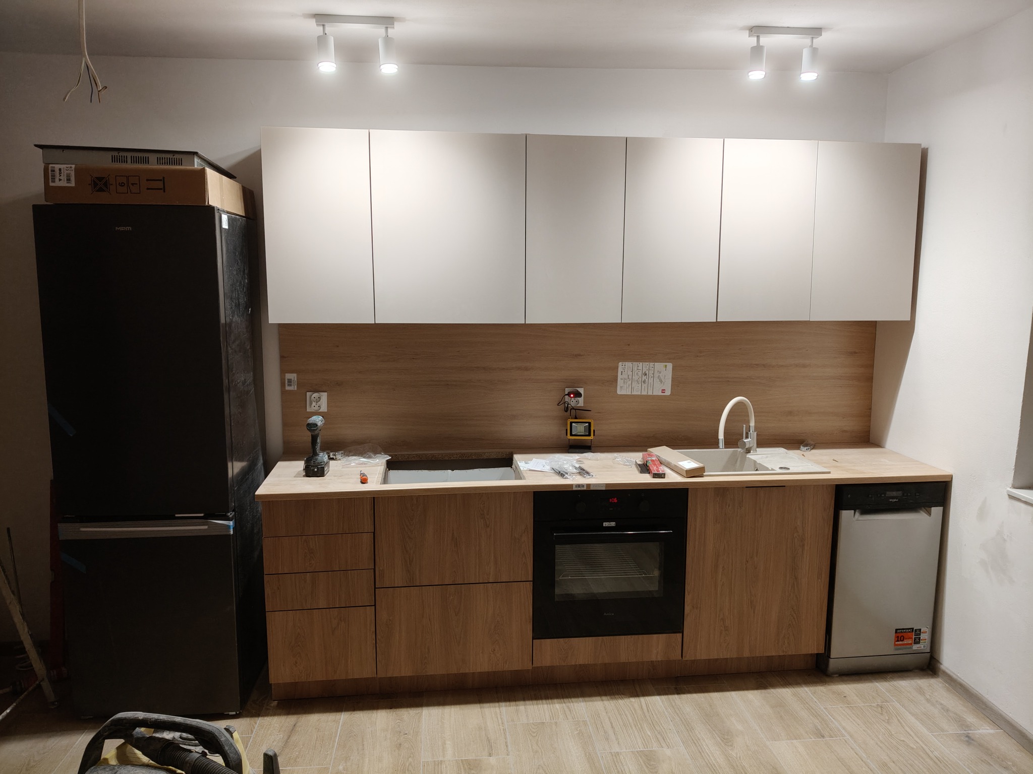 Nowoczesna kuchnia w trakcie montażu: białe szafki górne, drewnopodobne dolne, czarna lodówka z kartonem na górze, sprzęt AGD w zabudowie, widoczne narzędzia.