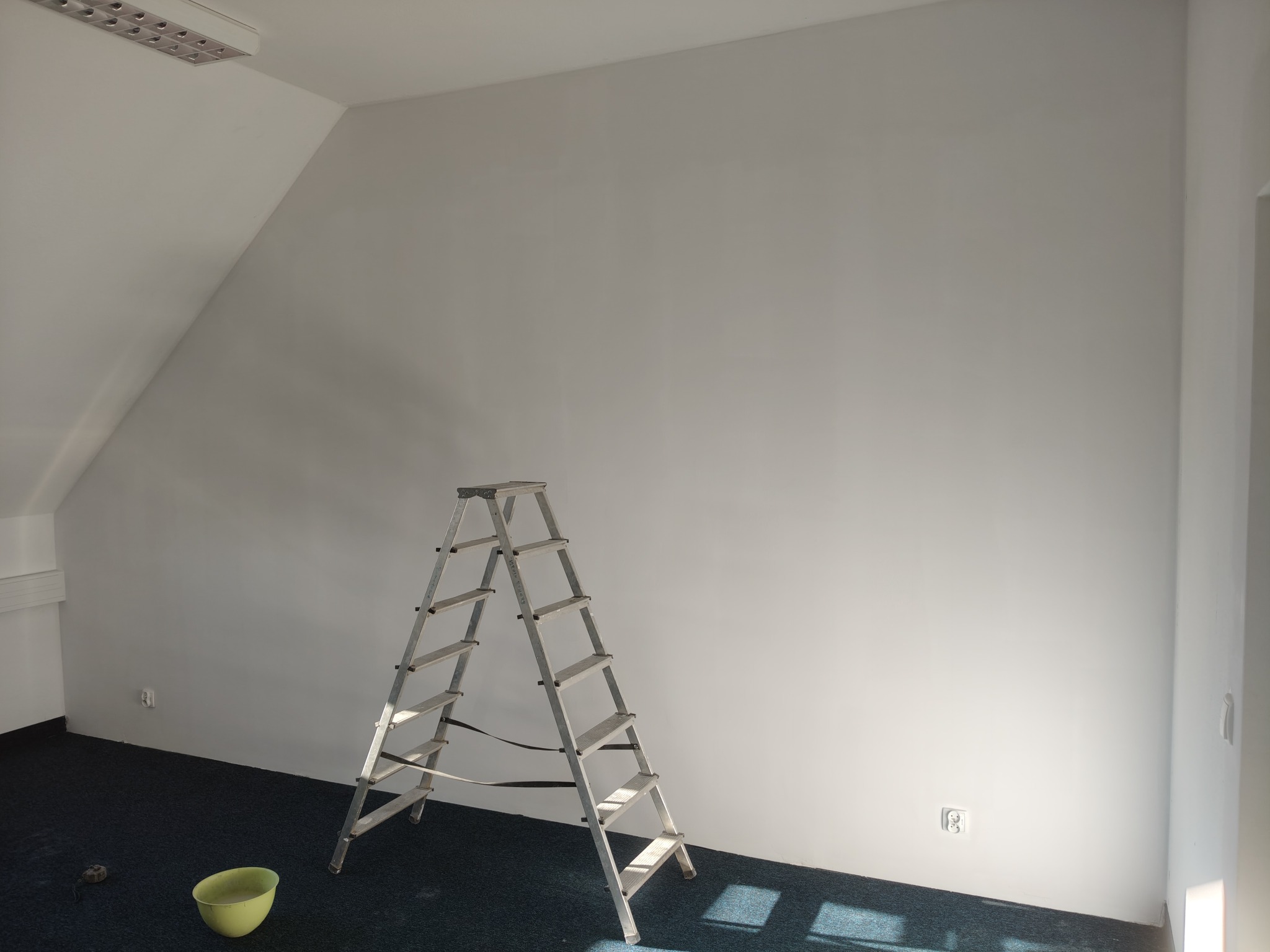 Wnętrze po malowaniu: białe ściany w pokoju na poddaszu, aluminiowa drabina, wiadro z farbą i niebieska wykładzina. Widoczne gniazdko elektryczne.
