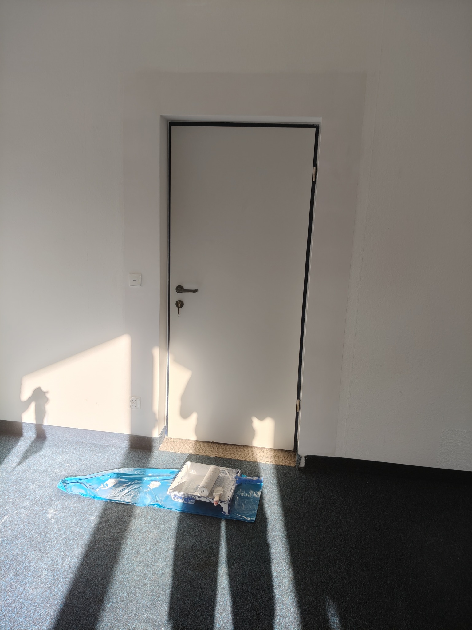 Białe drzwi z czarną ramą w nowym pomieszczeniu. Na podłodze zestaw malarski w folii ochronnej, rzucający cień na ścianę. Przygotowanie do malowania.