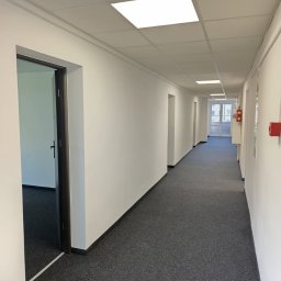 IKPM - Długi, pusty korytarz biurowy z szarą wykładziną, białymi ścianami i kwadratowymi lampami sufitowymi. Widoczne drzwi i system przeciwpożarowy.