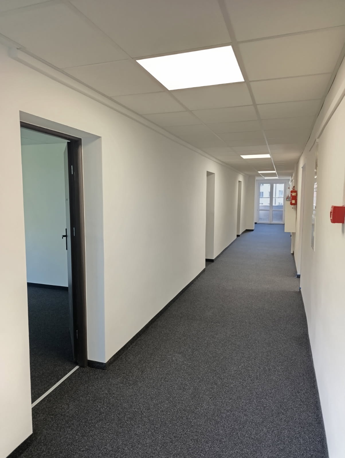 Długi, pusty korytarz biurowy z szarą wykładziną, białymi ścianami i kwadratowymi lampami sufitowymi. Widoczne drzwi i system przeciwpożarowy.