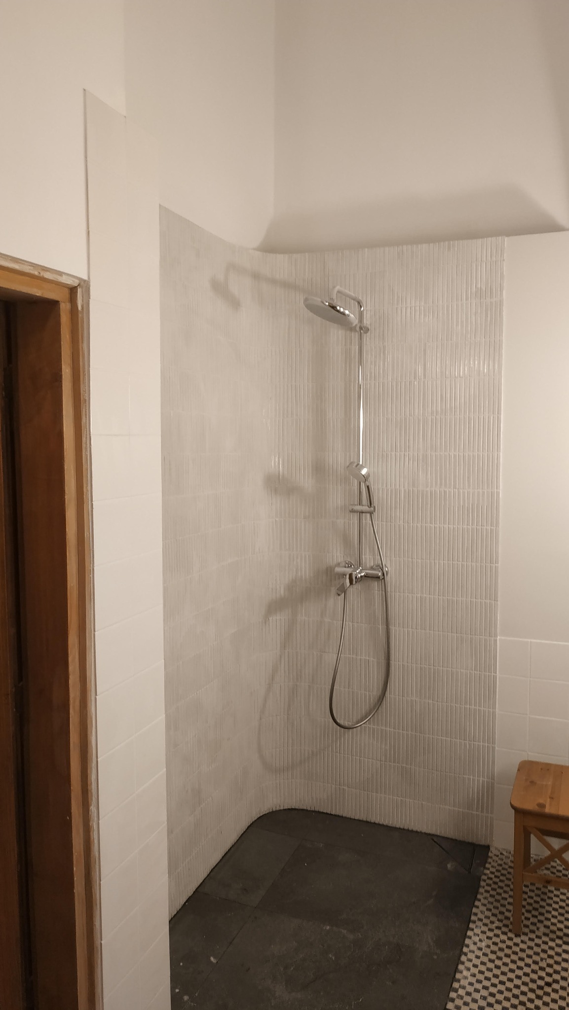 Wykończona kabina prysznicowa z chromowaną armaturą i białymi, pionowo ułożonymi płytkami. W tle widoczny fragment drewnianego stołka i wzorzystej podłogi.