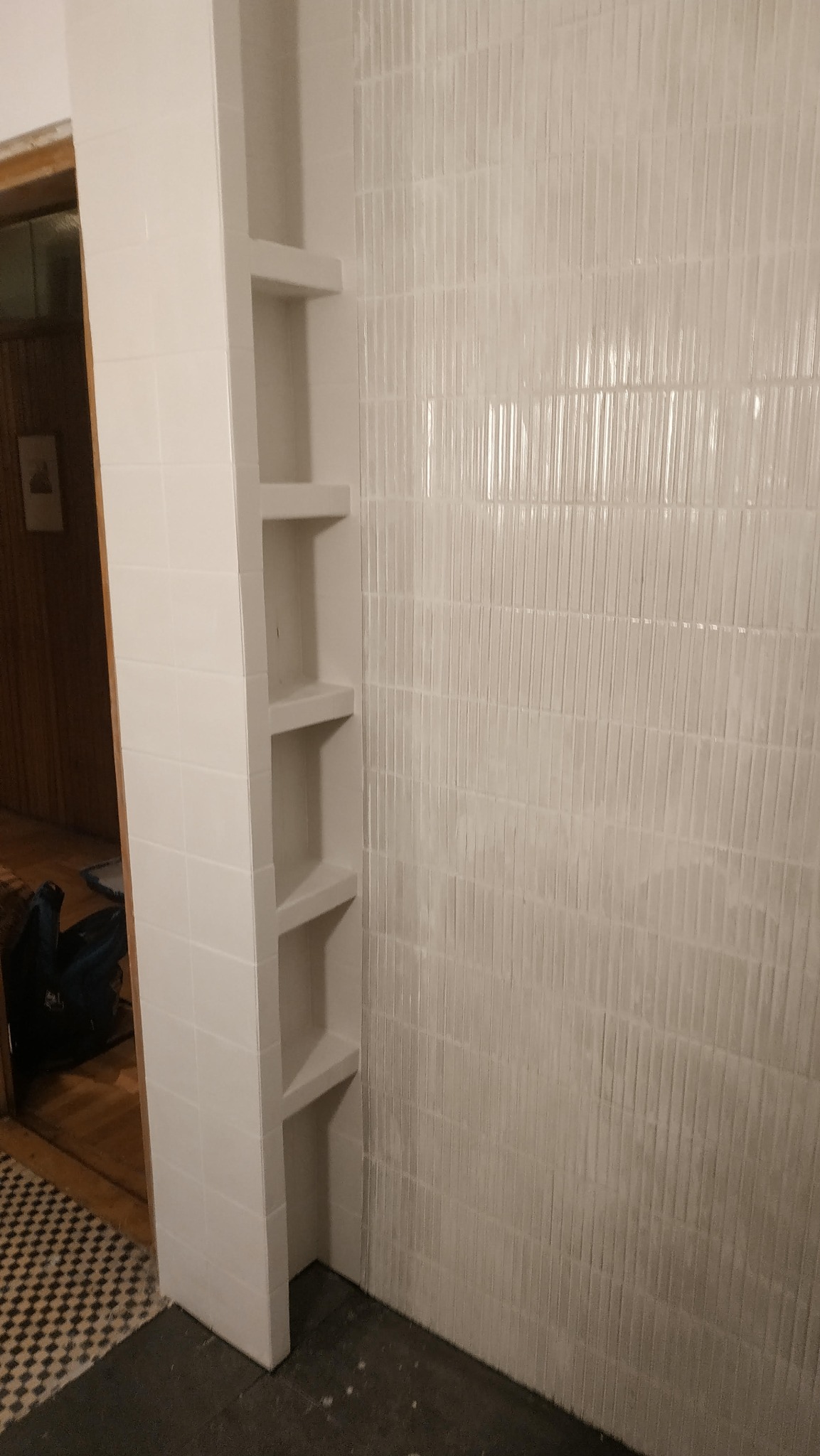 Biała łazienka po remoncie: pionowe płytki na ścianie i wnęka z półkami z płytek. Widoczny fragment podłogi i przejście do innego pomieszczenia.