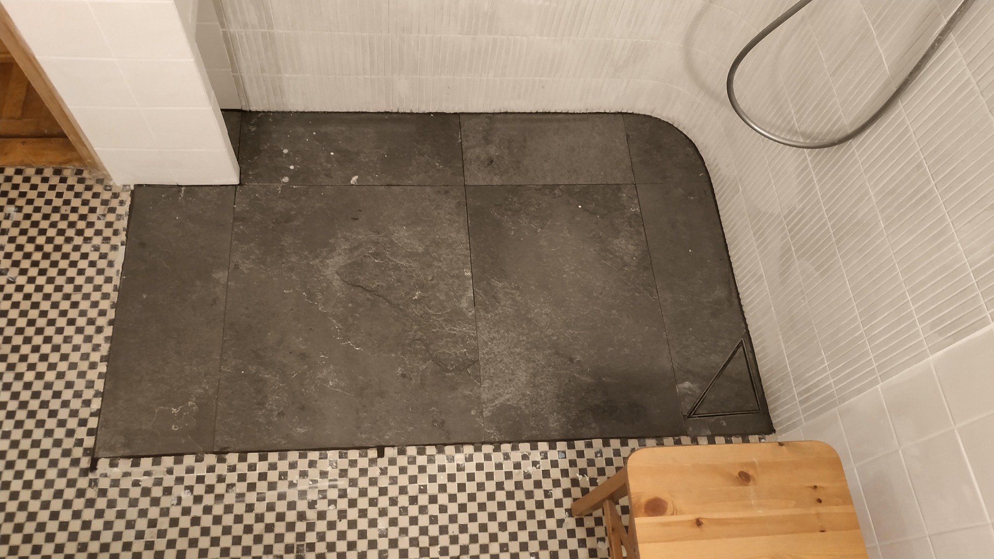 Wykończona strefa prysznicowa z ciemną, kamienną posadzką, odpływem liniowym i białymi, poziomymi płytkami na ścianach. Fragment drewnianego stołka w rogu.