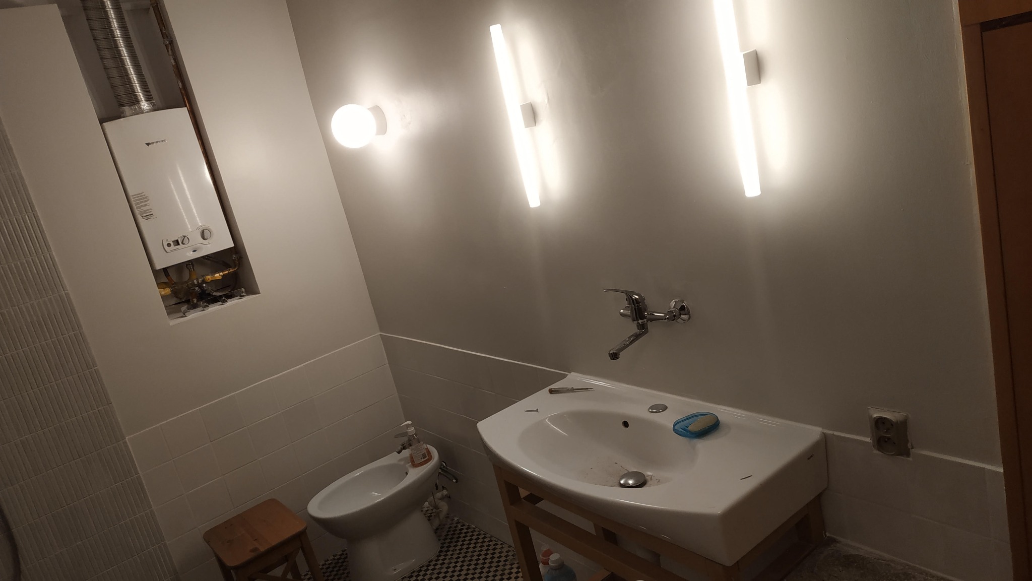 Wykończona łazienka z białymi płytkami, wiszącą umywalką na drewnianym stelażu, bidetem i piecem gazowym w zabudowie. Oświetlenie stanowią podłużne lampy LED.