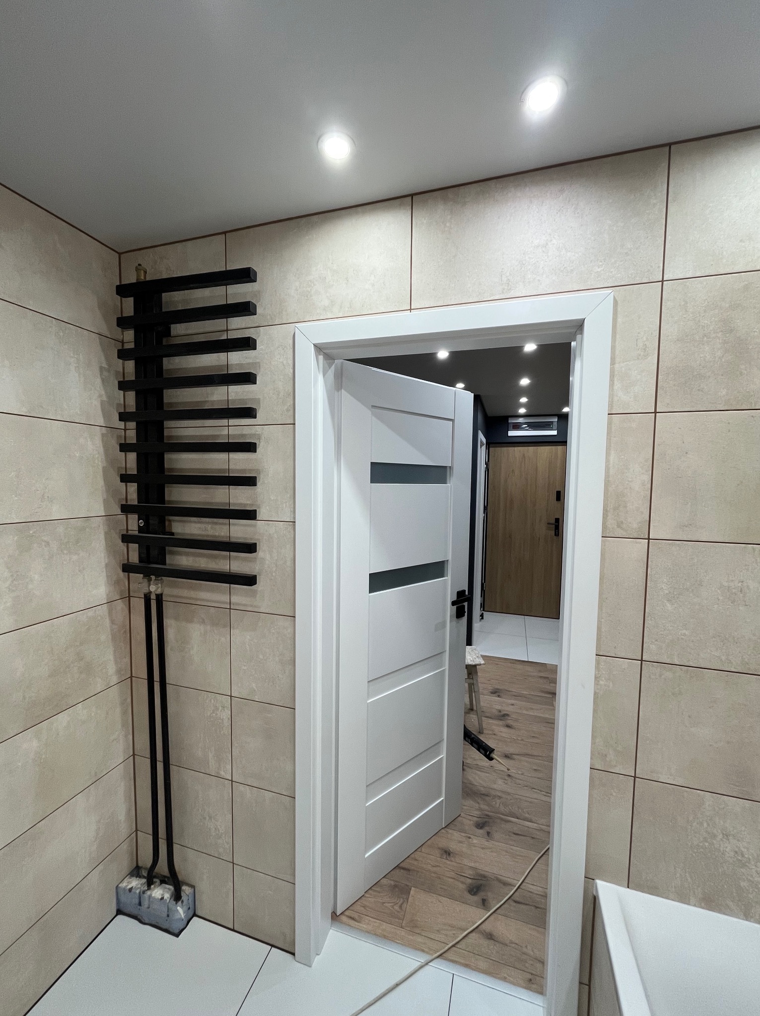 Wykończona łazienka w Krakowie: beżowe płytki, białe drzwi z przeszkleniami i czarny grzejnik drabinkowy. Widok na korytarz z drewnianą podłogą.