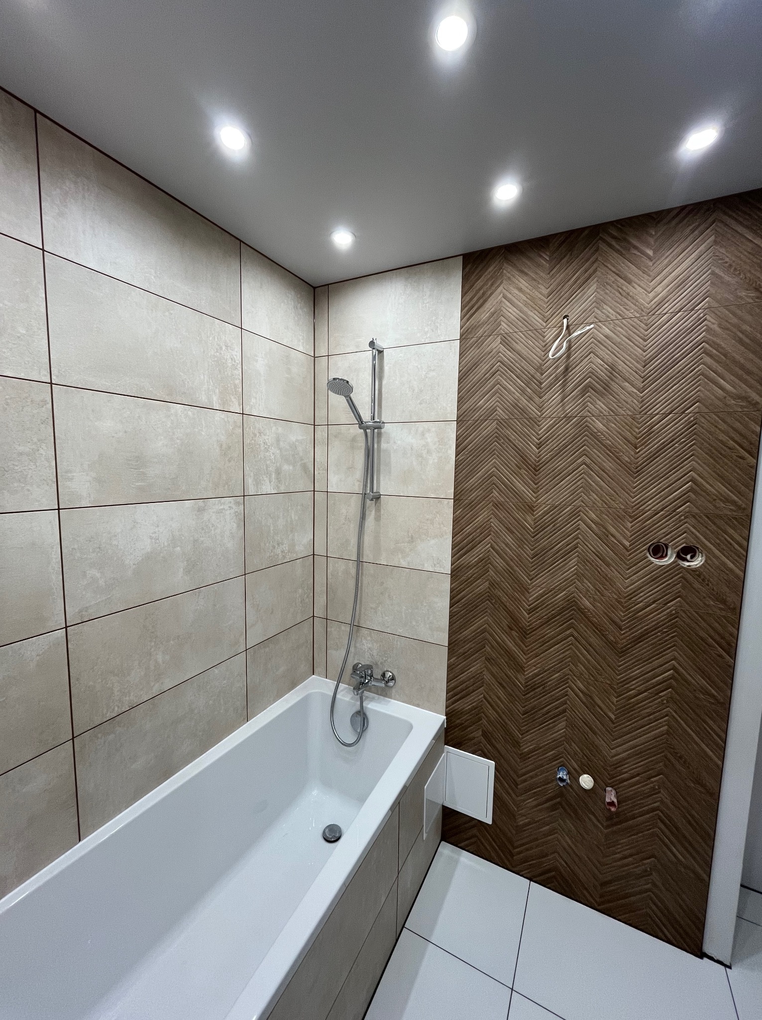 Nowoczesna łazienka po remoncie: biała wanna, beżowe płytki i dekoracyjna ściana z drewnopodobnych paneli ułożonych w jodełkę. Widoczne punkty świetlne w suficie.