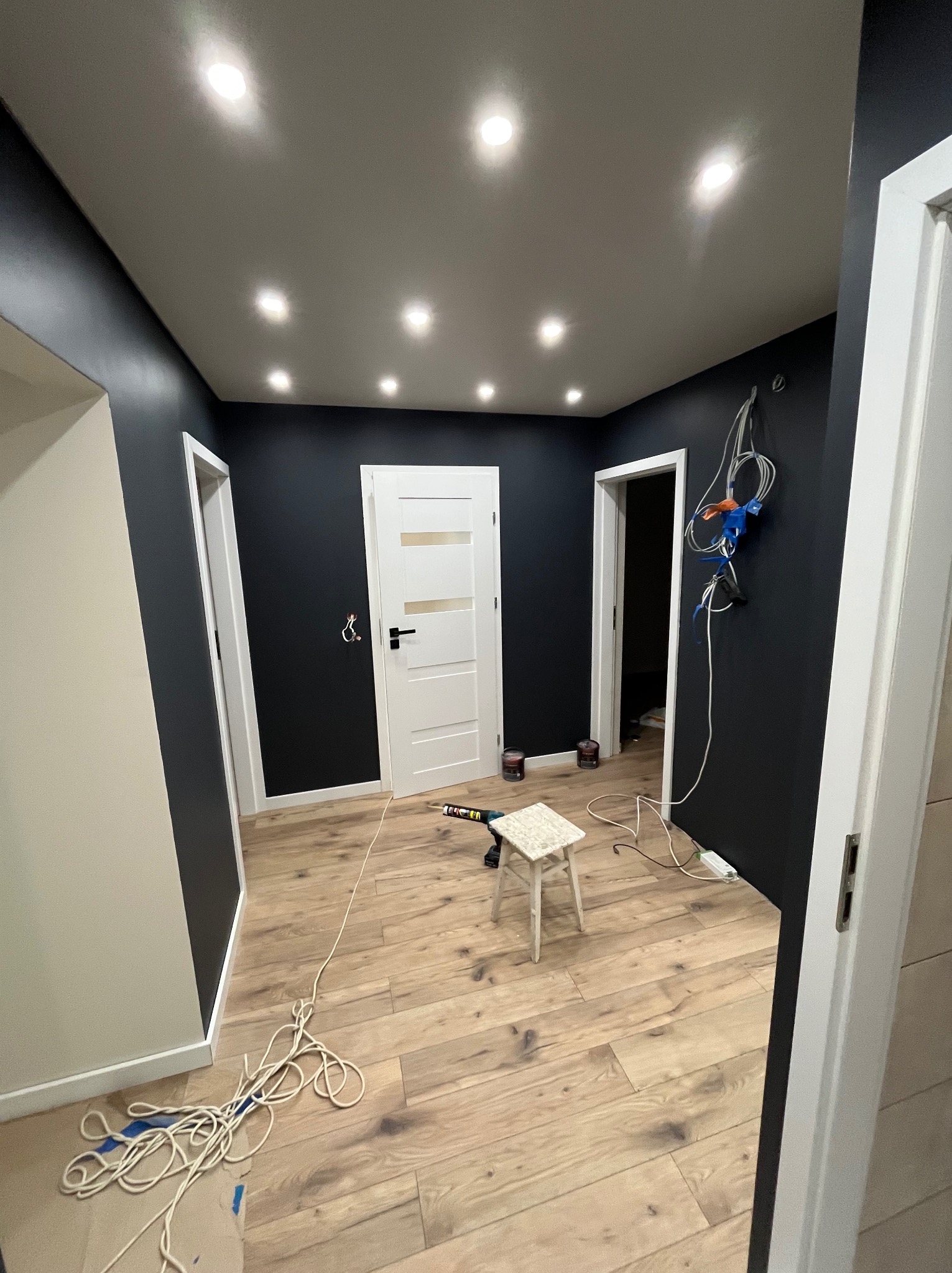 Wnętrze po malowaniu na ciemny grafit, z drewnianą podłogą i białymi drzwiami. Widoczne narzędzia malarskie i oświetlenie sufitowe.