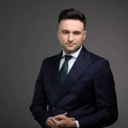 Portret mężczyzny w eleganckim, dwurzędowym garniturze w kratę, z zielonym krawatem, na gładkim, szarym tle. Dłonie splecione, stonowana kolorystyka, profesjonalny wygląd.
