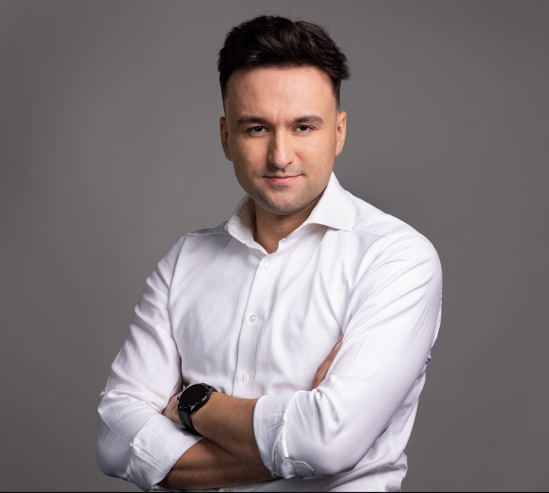 Portret mężczyzny w białej koszuli z zegarkiem na tle szarego studia. Uśmiechnięty, pewny siebie, z założonymi rękami. Profesjonalny wygląd, dobre oświetlenie.