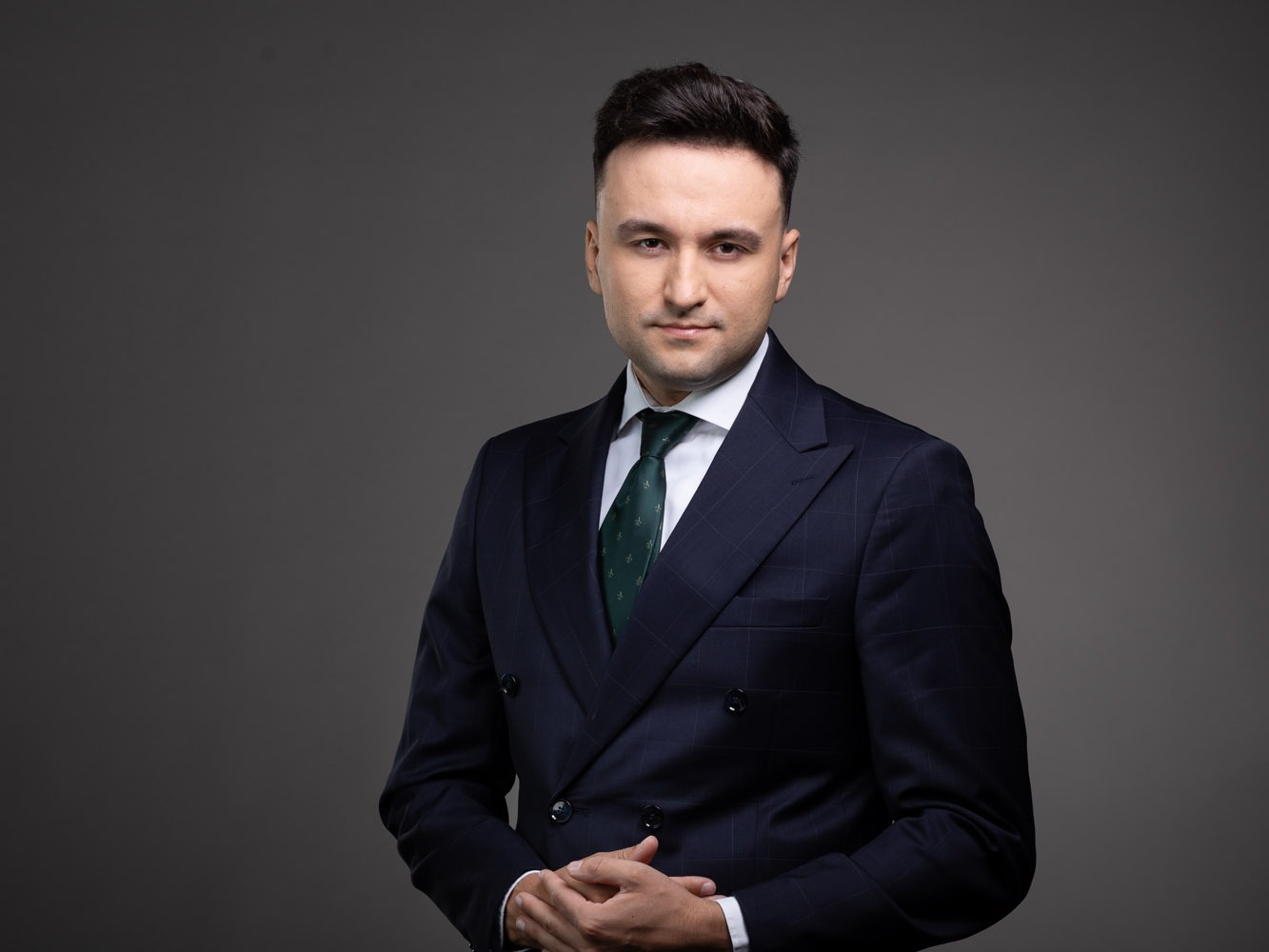 Portret mężczyzny w eleganckim, dwurzędowym garniturze w kratę, z zielonym krawatem, na gładkim, szarym tle. Dłonie splecione, stonowana kolorystyka, profesjonalny wygląd.