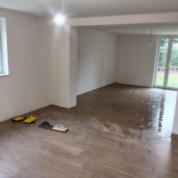 ADMAGBUD ADAM KNAP - Układanie płytek podłogowych imitujących drewno w jasnym, pustym pomieszczeniu. Widoczne narzędzia glazurnicze i świeżo ułożona, mokra powierzchnia.