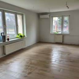 ADMAGBUD ADAM KNAP - Jasne, przestronne wnętrze po remoncie: drewnopodobna podłoga, świeżo pomalowane ściany, dwa okna z widokiem na zieleń. Widoczny grzejnik i klimatyzator.