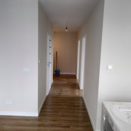 ADMAGBUD ADAM KNAP - Jasny korytarz po remoncie z panelami podłogowymi, świeżo pomalowanymi ścianami i nowymi drzwiami. W głębi widoczne wiadro i mop.
