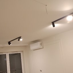 ADMAGBUD ADAM KNAP - Nowoczesne oświetlenie sufitowe z reflektorami na szynoprzewodzie w jasnym wnętrzu. Widoczny klimatyzator i okno. Minimalistyczny design.