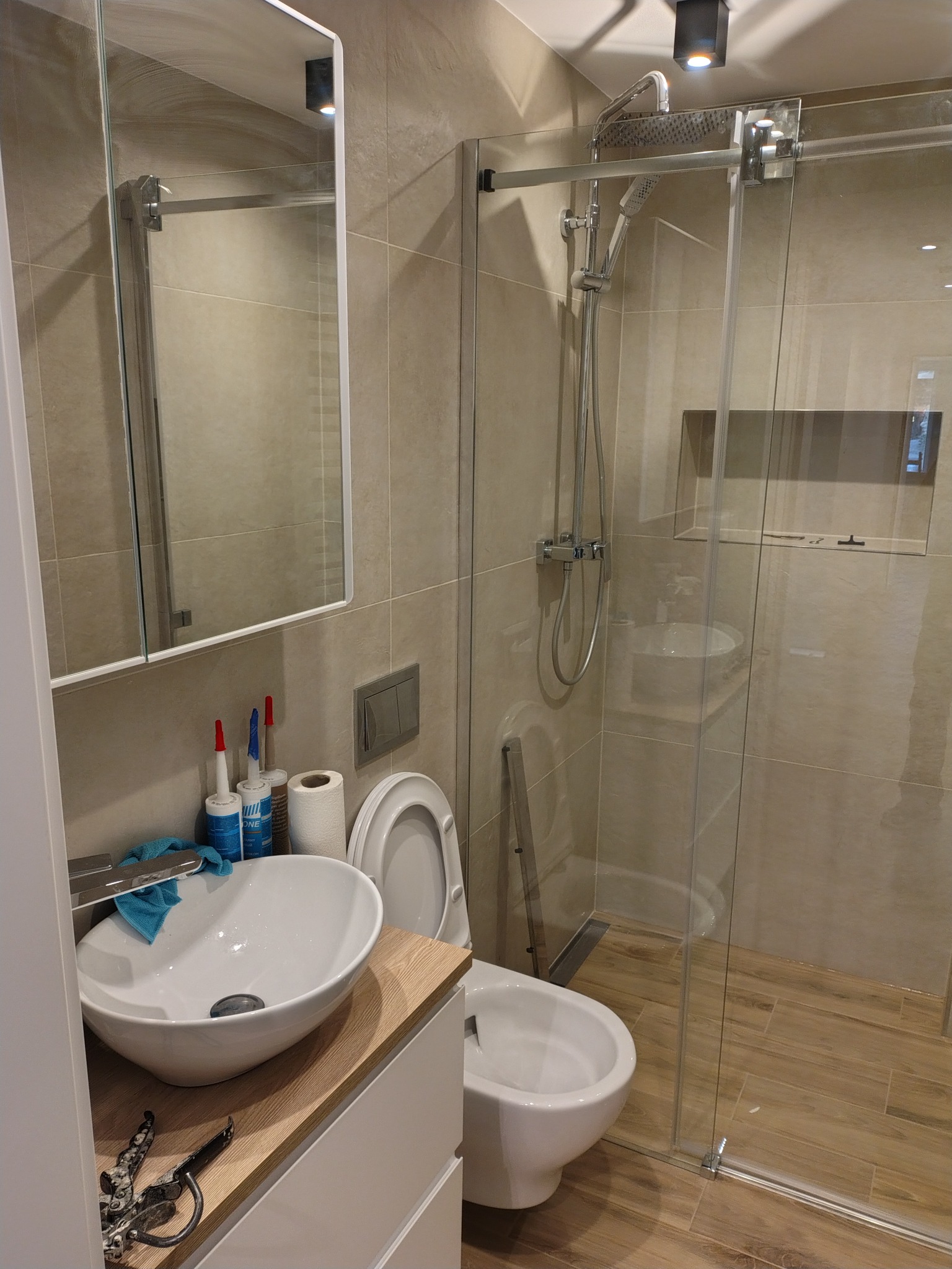 Nowoczesna łazienka z kabiną prysznicową typu walk-in, umywalką nablatową i bidetem. Beżowe płytki imitujące drewno, minimalistyczne oświetlenie punktowe.