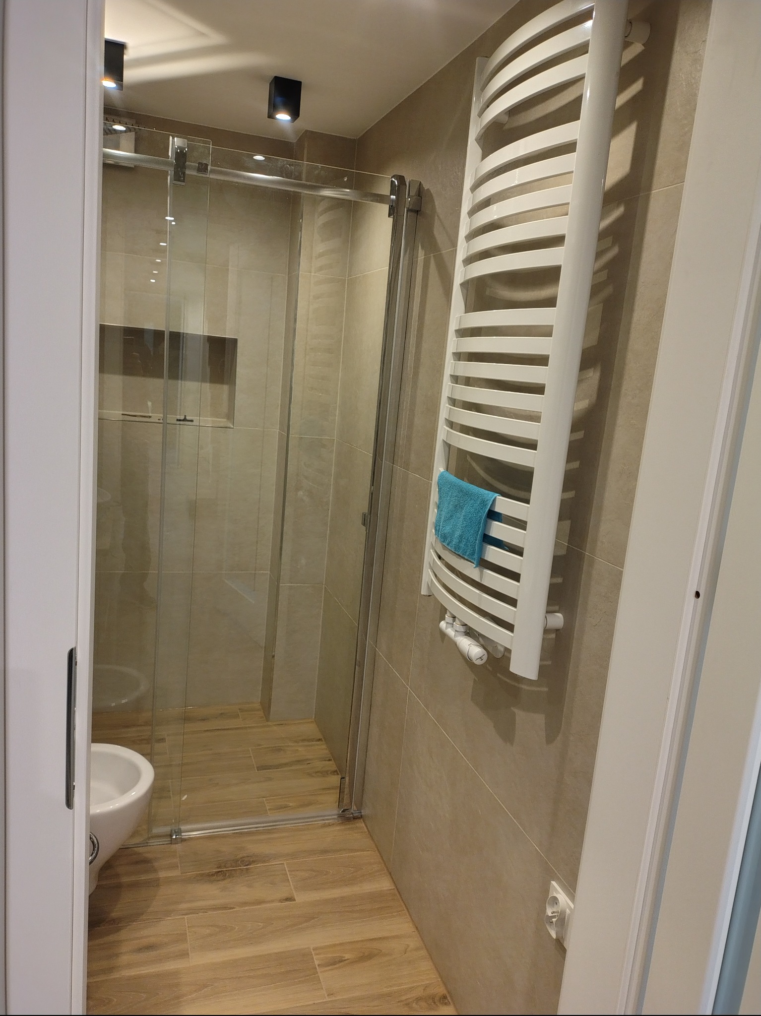 Nowoczesna łazienka z kabiną prysznicową ze szklanymi drzwiami przesuwnymi, grzejnikiem drabinkowym z ręcznikiem i drewnopodobną podłogą. Minimalistyczny design.