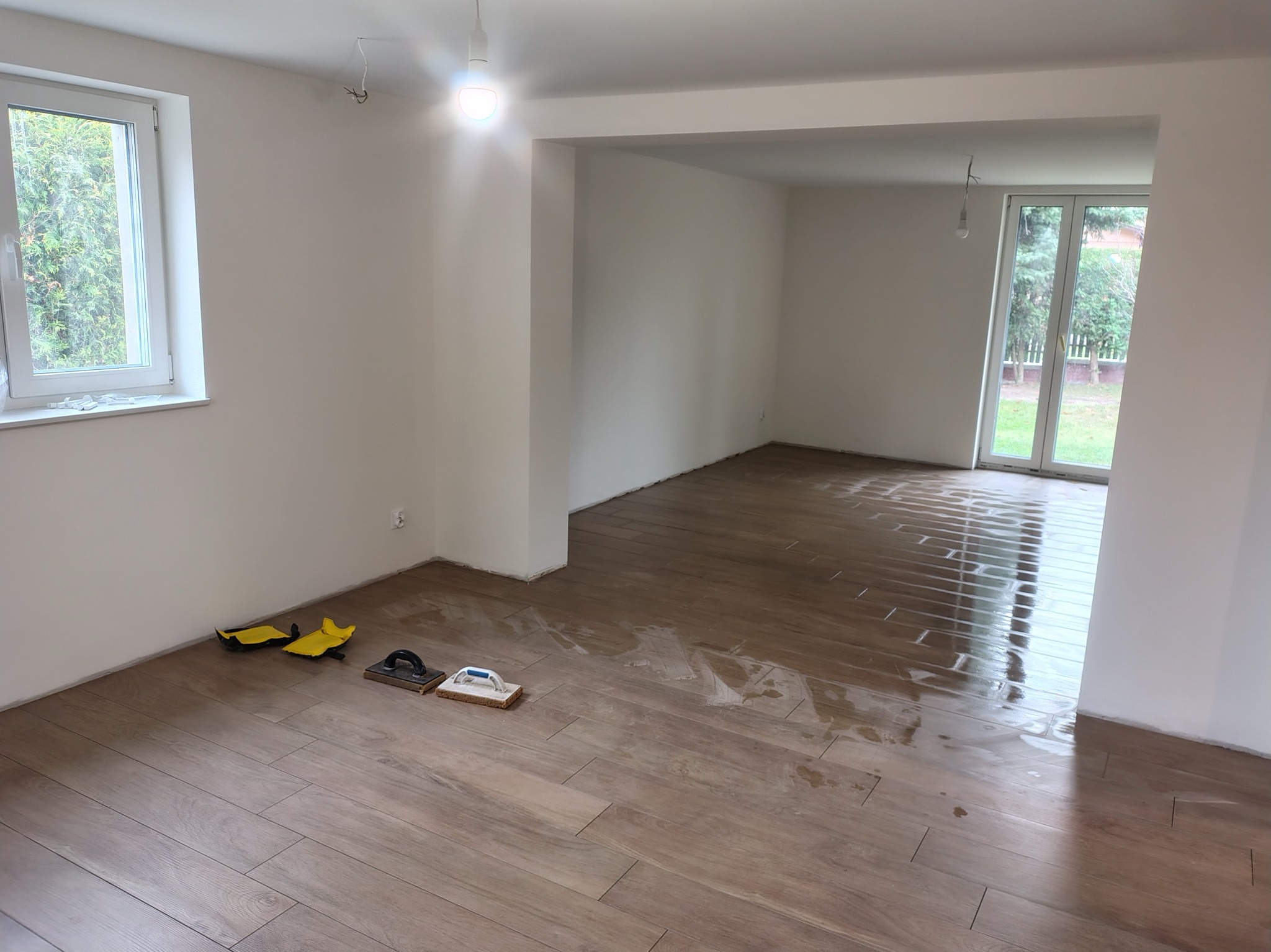 Układanie płytek podłogowych imitujących drewno w jasnym, pustym pomieszczeniu. Widoczne narzędzia glazurnicze i świeżo ułożona, mokra powierzchnia.