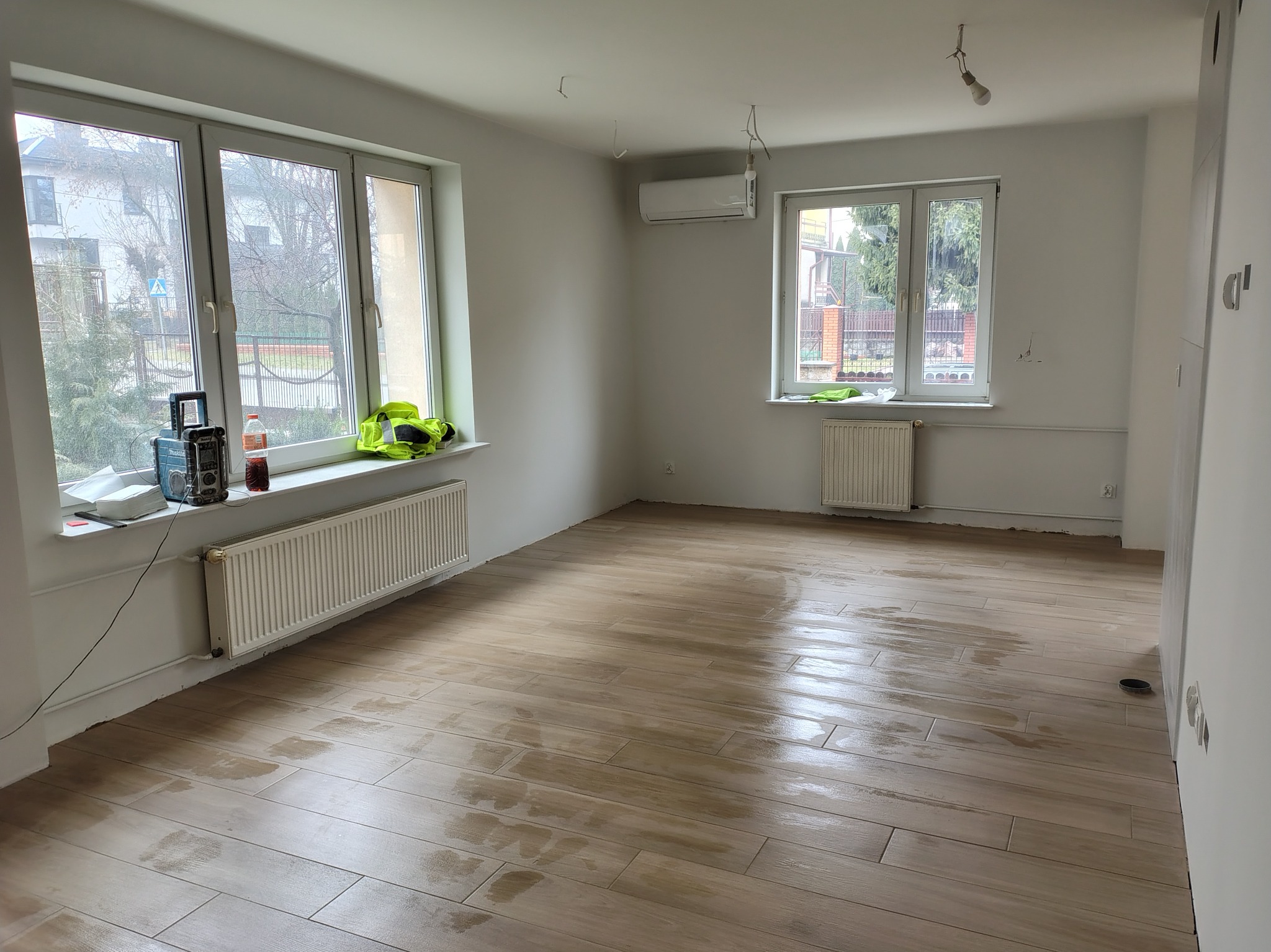 Jasne, przestronne wnętrze po remoncie: drewnopodobna podłoga, świeżo pomalowane ściany, dwa okna z widokiem na zieleń. Widoczny grzejnik i klimatyzator.