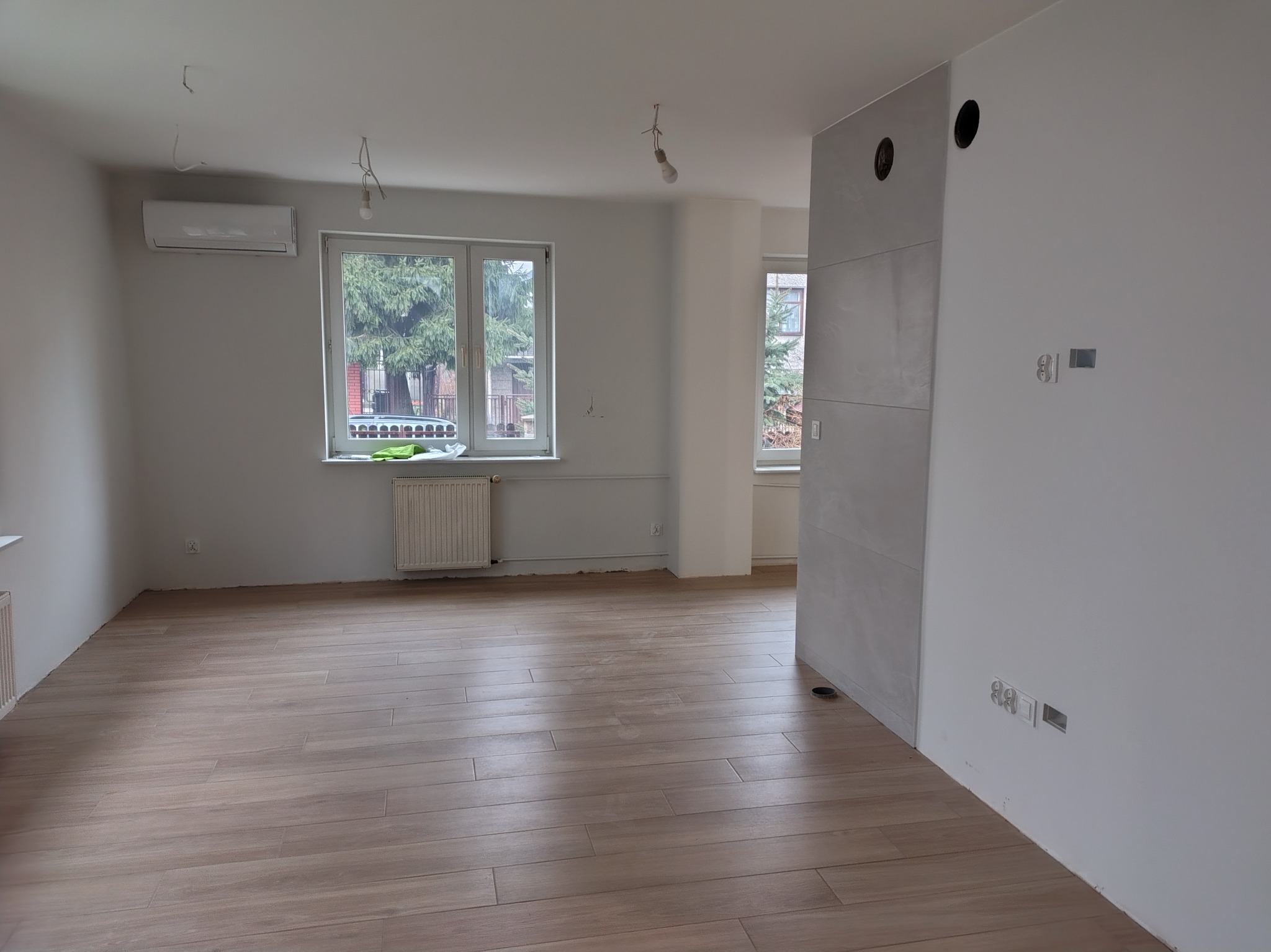 Jasne, przestronne wnętrze po remoncie: drewnopodobna podłoga, białe ściany z gniazdkami, grzejnik pod oknem. Widoczne niedokończone punkty oświetleniowe.