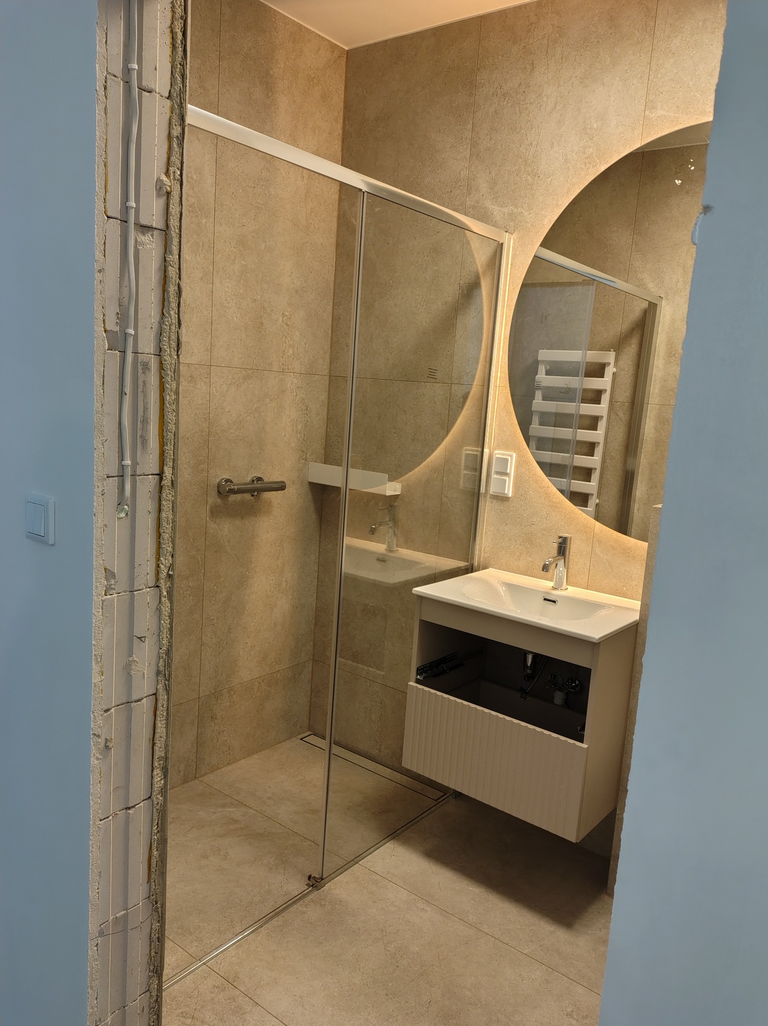 Beżowa łazienka z kabiną prysznicową ze szklanymi drzwiami, umywalką z szafką i okrągłym lustrem z podświetleniem LED. Widoczny fragment ściany w stanie surowym.