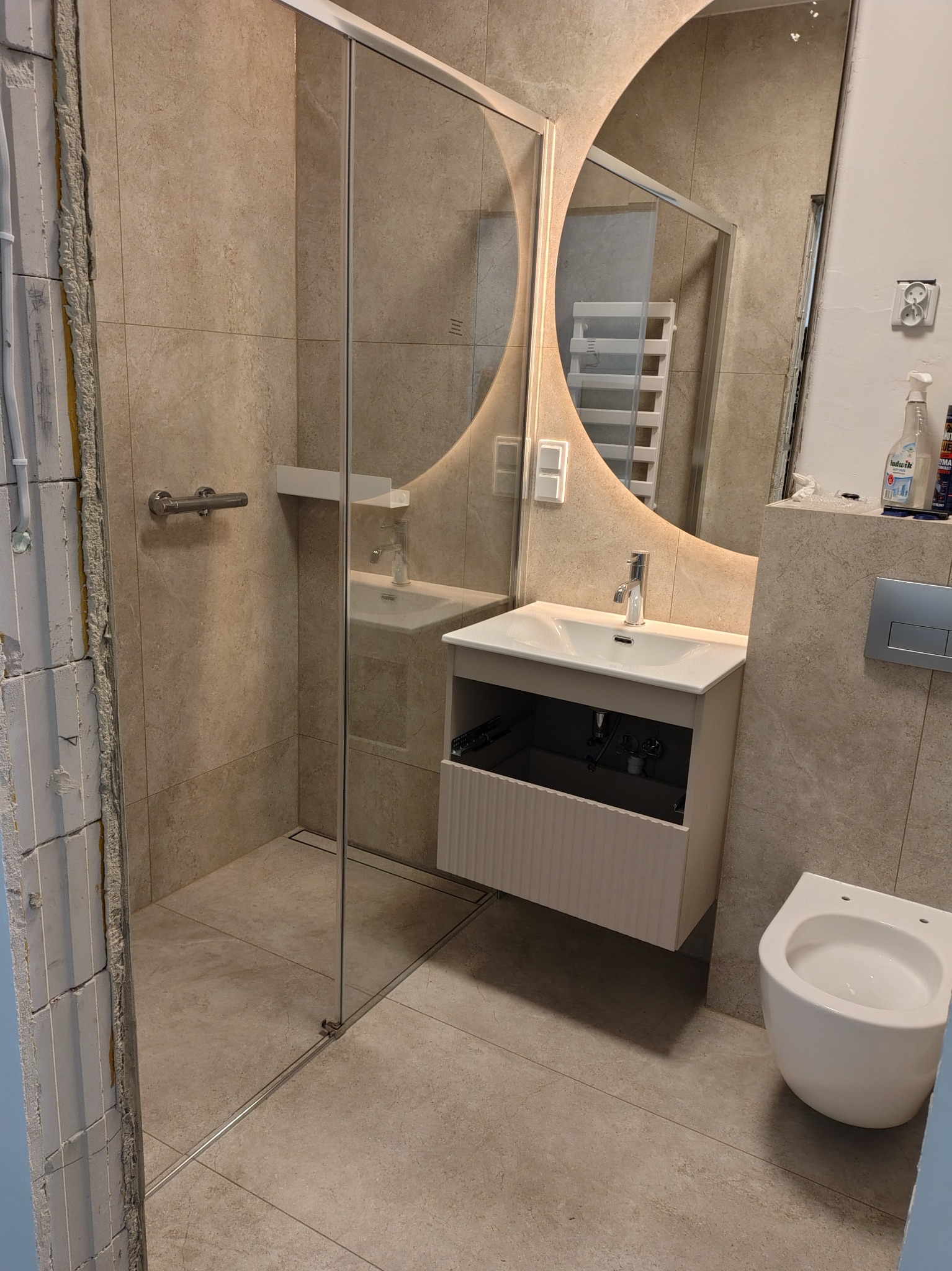 Nowoczesna łazienka w beżowych płytkach z kabiną prysznicową typu walk-in, umywalką z szafką i podświetlanym lustrem. Widoczne niedokończone wykończenia przy drzwiach.