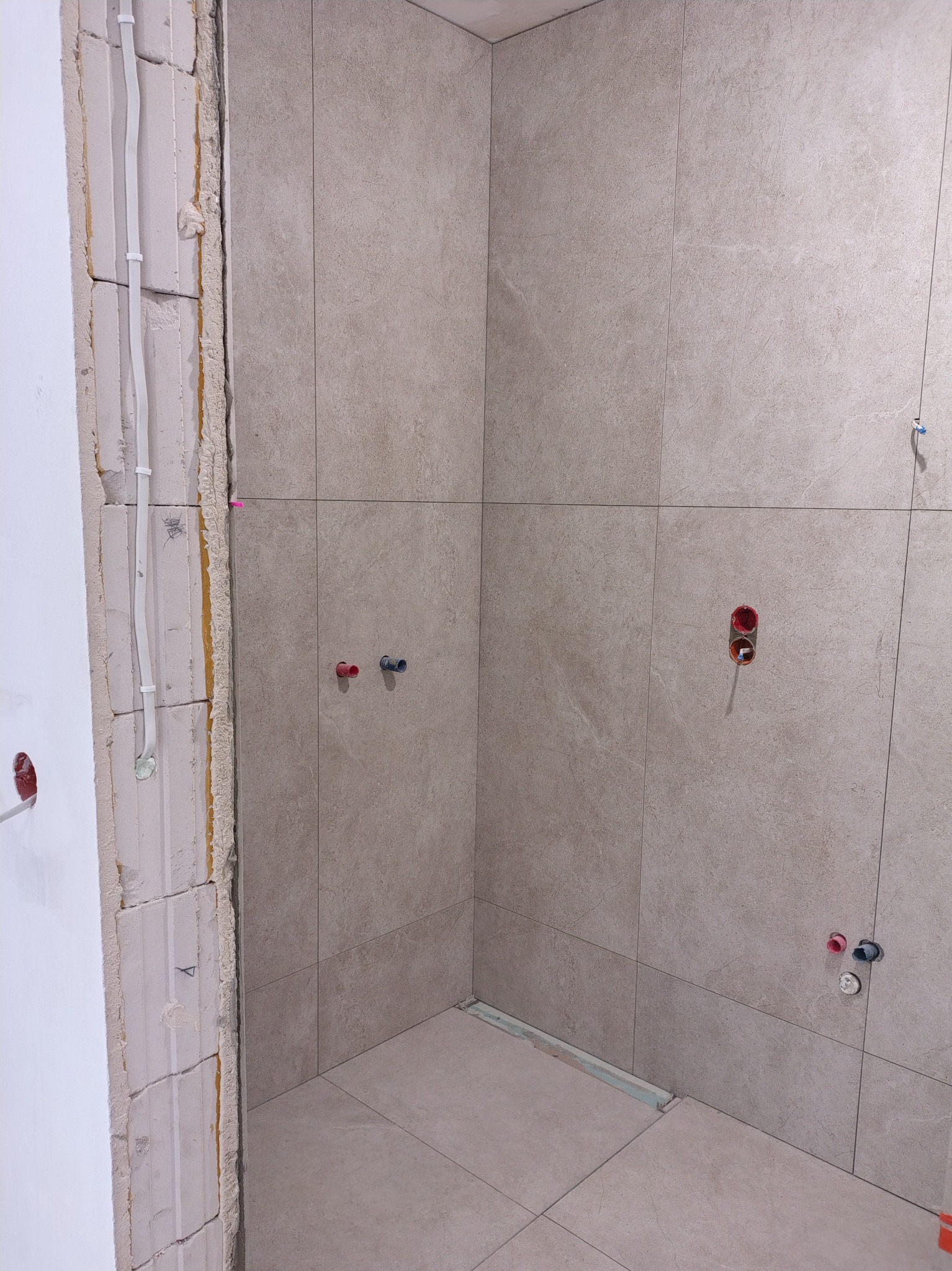 Wnętrze łazienki w trakcie wykańczania. Widoczne płytki na ścianach, przygotowane instalacje wodne i elektryczne, oraz surowy mur z instalacjami w peszlach.
