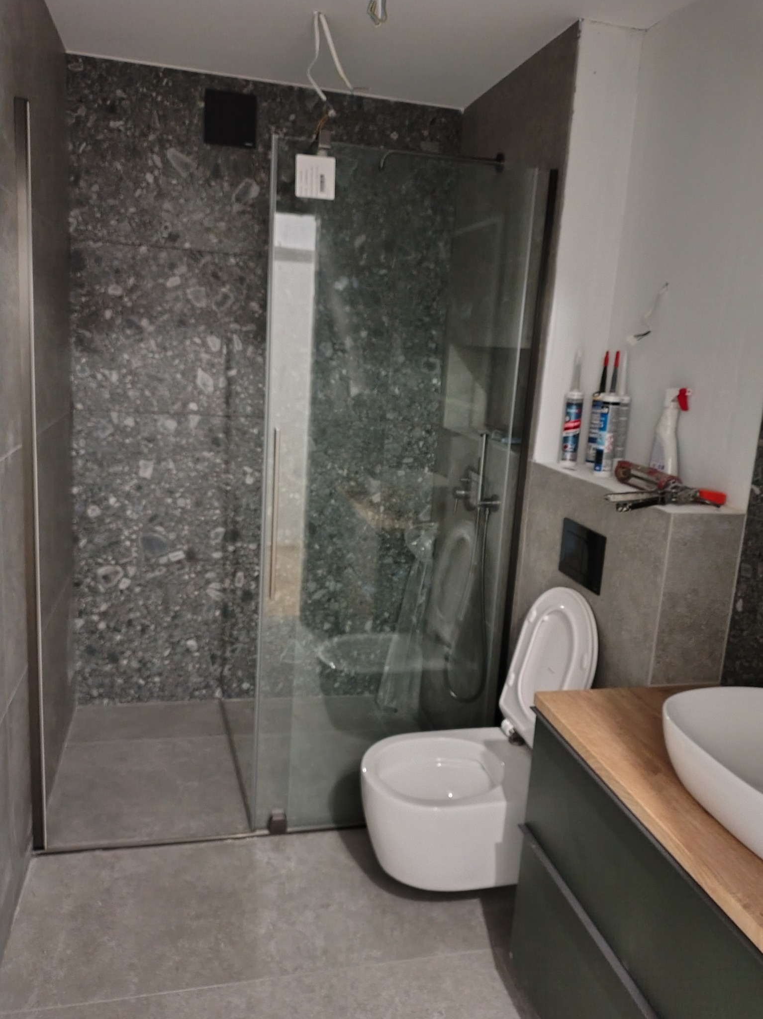 Nowoczesna łazienka po remoncie: kabina prysznicowa ze szklanymi drzwiami, podwieszana toaleta i umywalka na drewnianym blacie. Widoczne narzędzia i materiały budowlane.