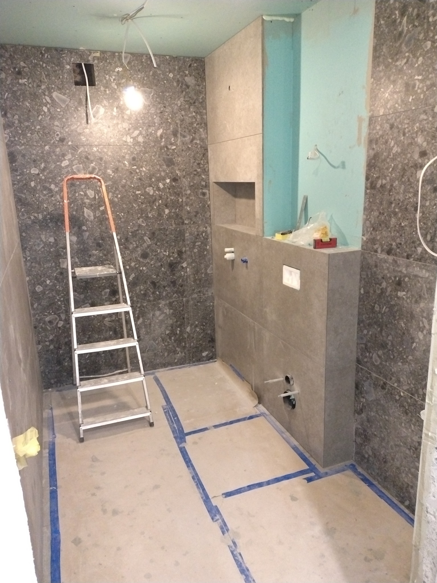 Łazienka w trakcie prac wykończeniowych: szare płytki na ścianach, zabudowa WC, drabina malarska. Widoczne instalacje i zabezpieczenia podłogi. Oświetlenie sufitowe.