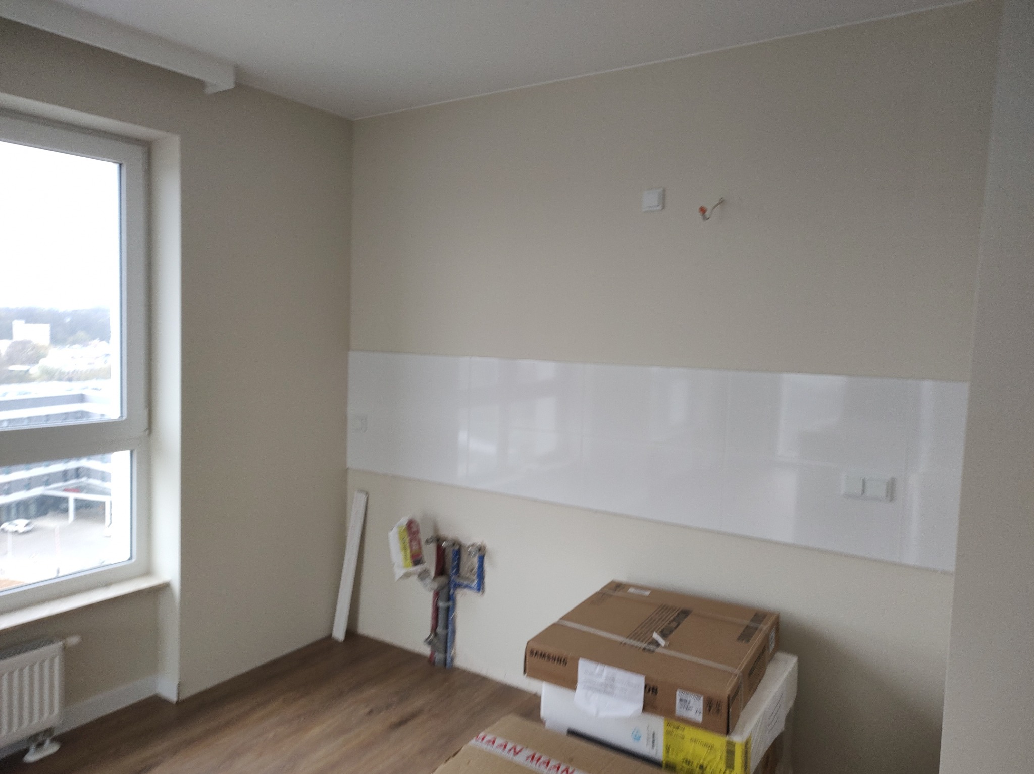 Wnętrze w trakcie wykańczania: białe panele ścienne, instalacja hydrauliczna, kartony z wyposażeniem. Widoczne drewniane panele podłogowe i okno z widokiem na miasto.