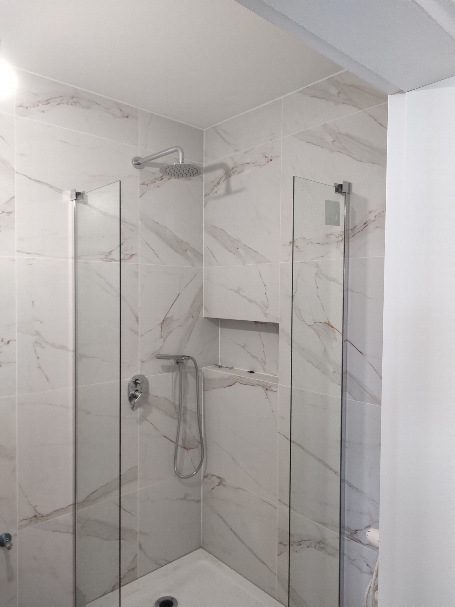 Nowoczesna kabina prysznicowa z marmurowymi płytkami, szklanymi ściankami i chromowaną armaturą. Wnęka w ścianie na kosmetyki. Minimalistyczny design.