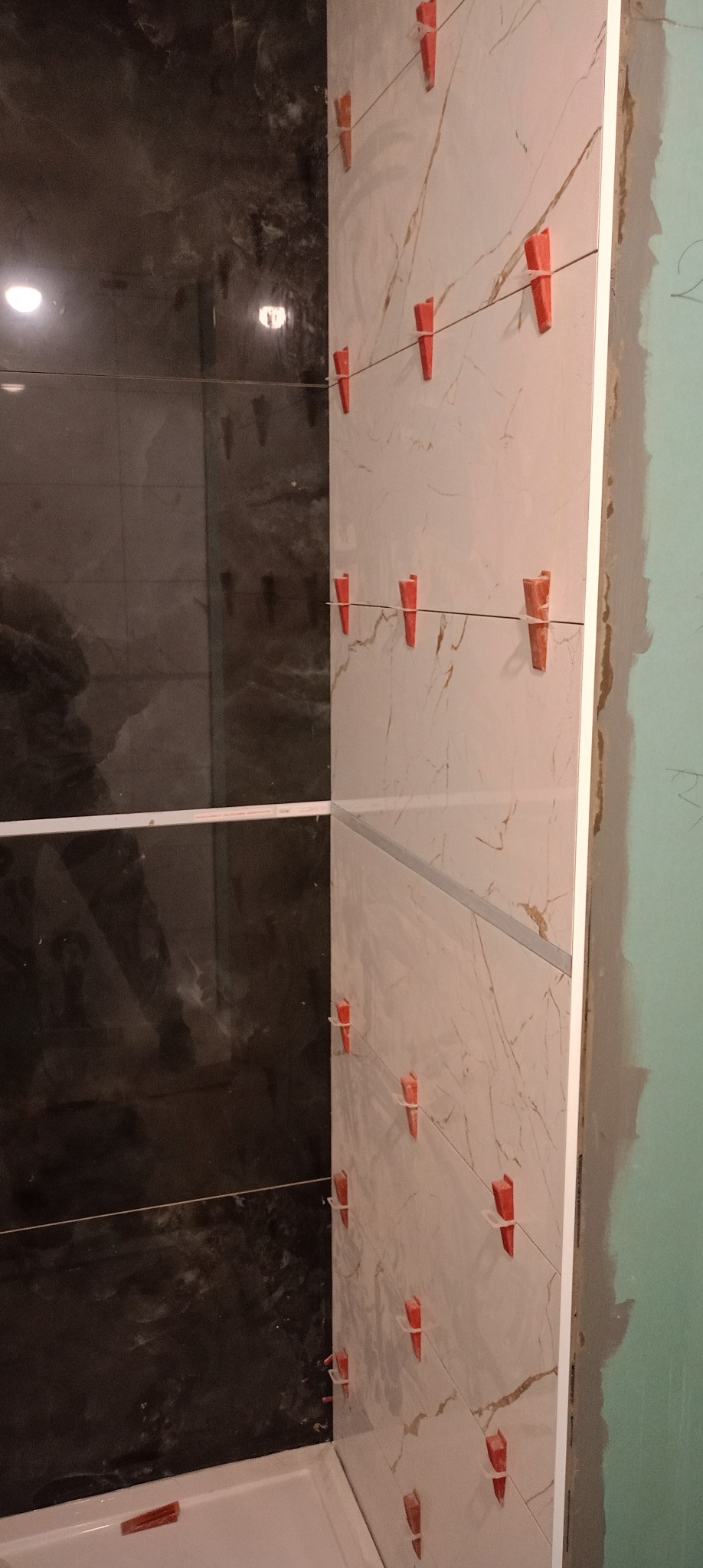 Układanie płytek marmurowych w łazience z użyciem klipsów poziomujących. Widoczna czarna ściana z płytek i fragment wanny. Etap prac wykończeniowych.