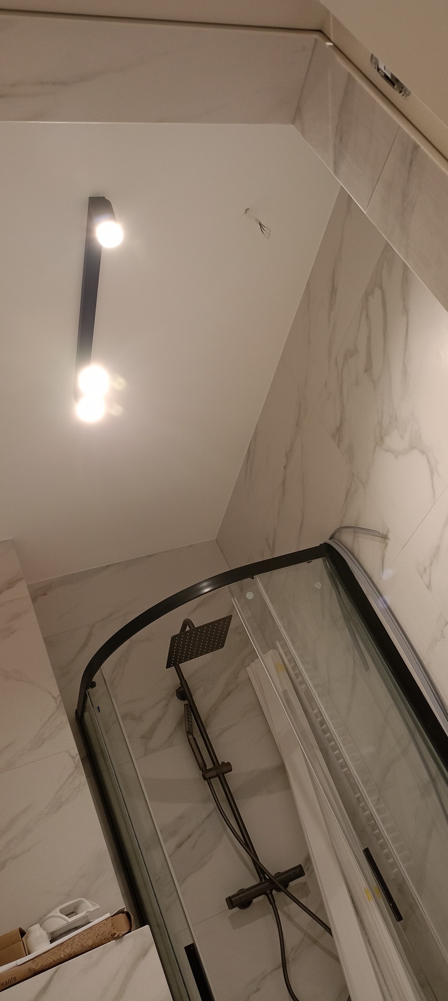 Nowoczesna kabina prysznicowa z czarną armaturą i deszczownicą, wykończona marmurowymi płytkami. Oświetlenie punktowe na suficie. Widok z dołu, podkreślający wysokość.