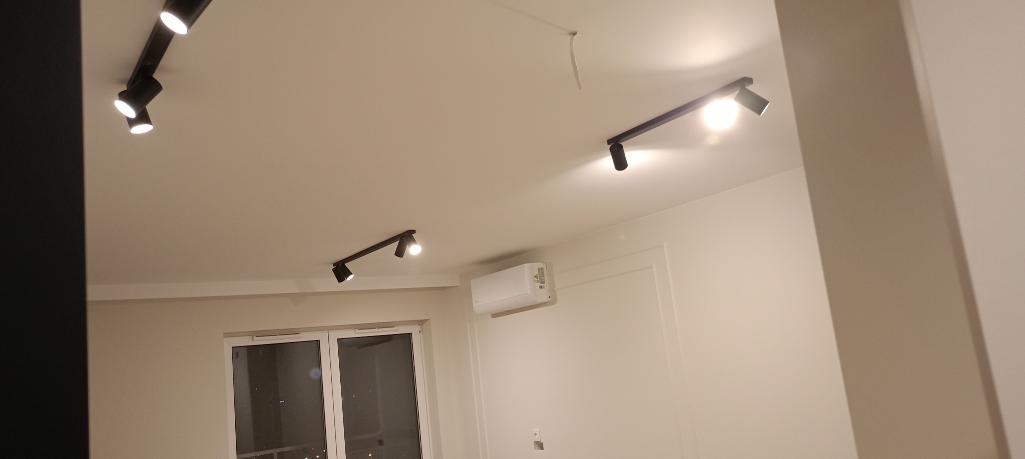 Nowoczesne oświetlenie sufitowe z reflektorami na szynoprzewodzie w jasnym wnętrzu. Widoczny klimatyzator i okno. Minimalistyczny design.