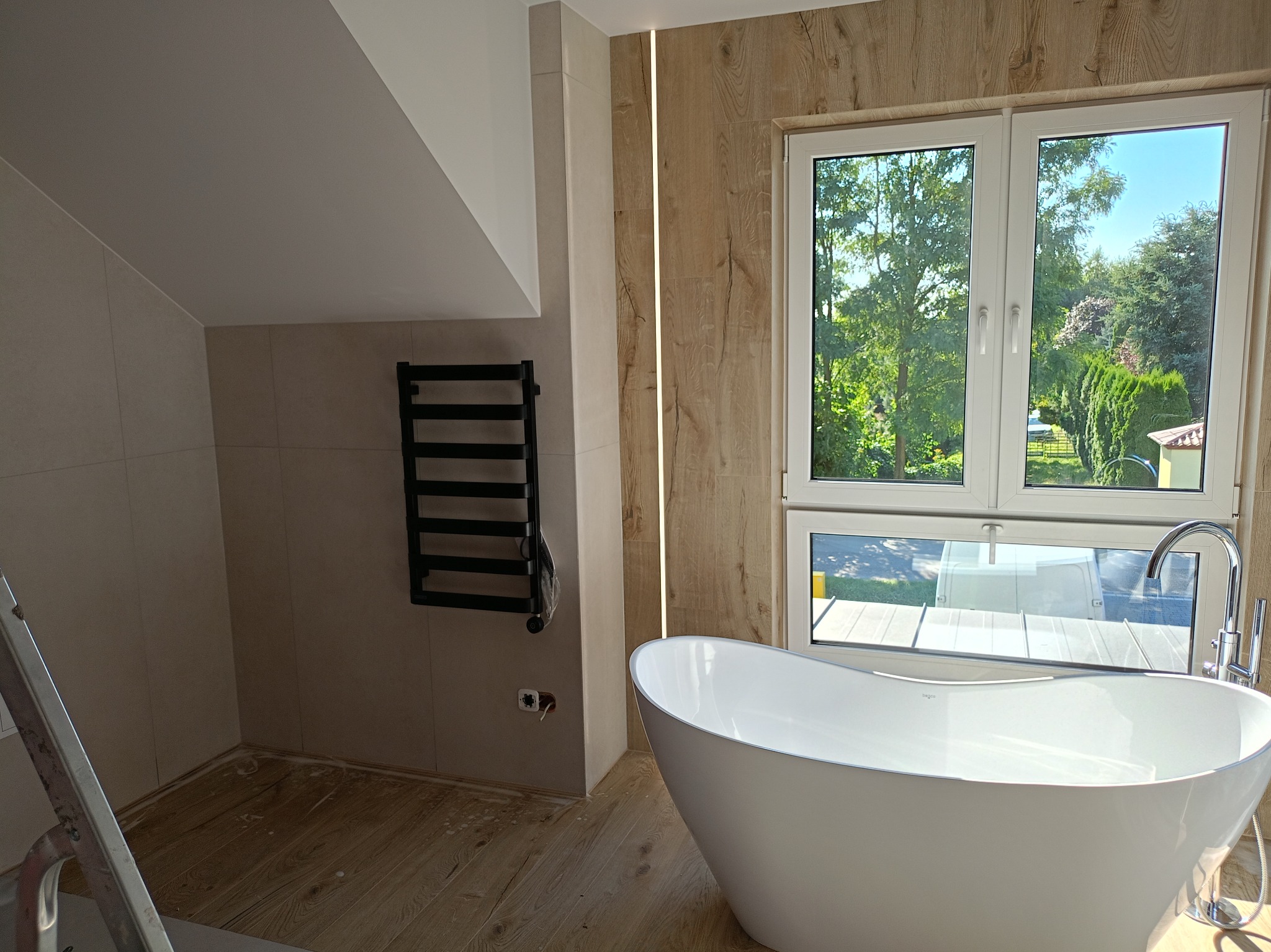 Nowoczesna łazienka z wolnostojącą wanną na tle okna z widokiem na zieleń. Na ścianie grzejnik drabinkowy i płytki imitujące drewno. Częściowo widoczna drabina.