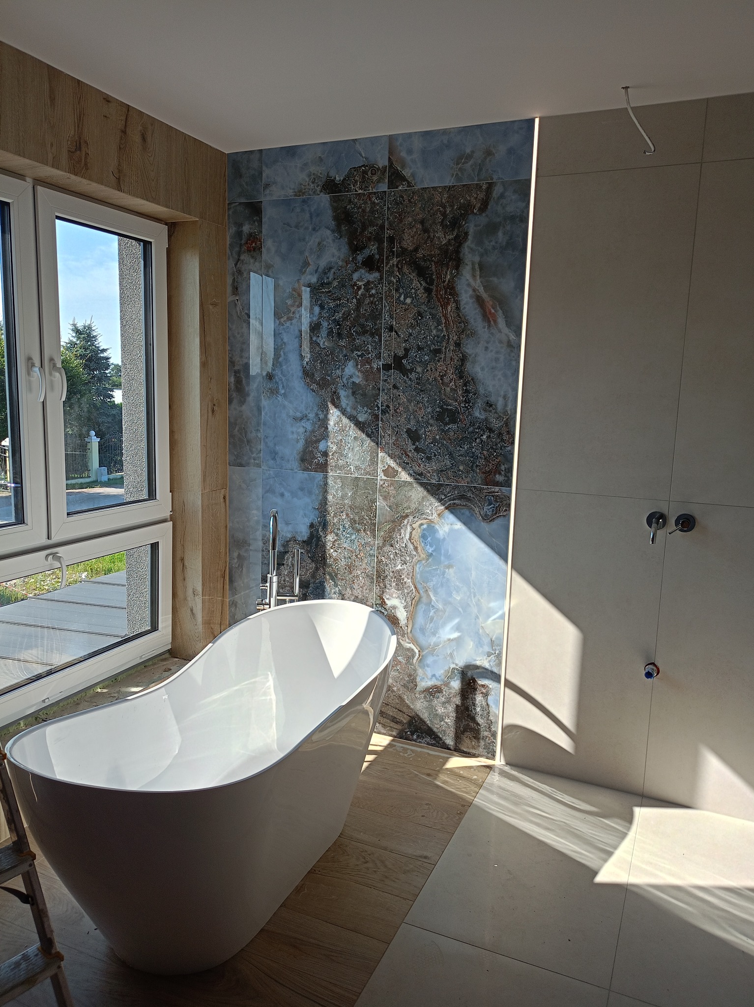 Nowoczesna łazienka z wolnostojącą wanną, marmurowymi płytkami i oknem. Widoczne elementy instalacji i drabina malarska, sugerujące prace wykończeniowe w toku.