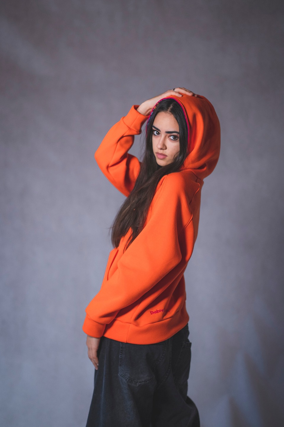 Modelka w pomarańczowej bluzie z kapturem i ciemnych jeansach pozuje na szarym tle, prezentując odzież.