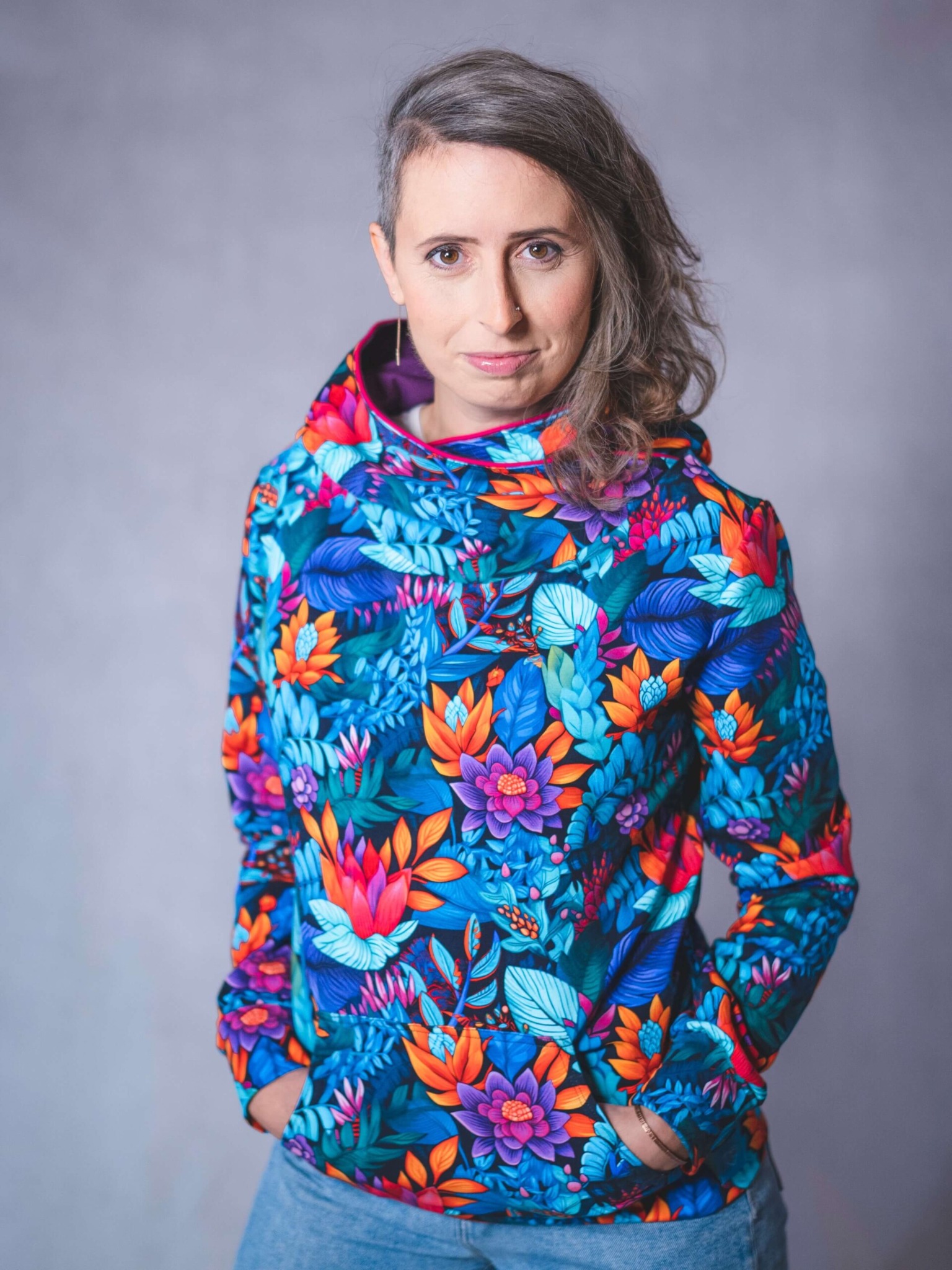 Kobieta w bluzie z kapturem, z barwnym, kwiatowym wzorem, pozuje na neutralnym tle. Widoczny fragment jeansów i ręce w kieszeniach.