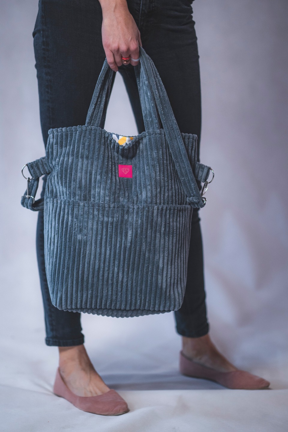 Szara, sztruksowa torba shopper z różową naszywką w kształcie serca, trzymana przez osobę w czarnych jeansach i różowych balerinach. Widoczny wzór wewnątrz torby.
