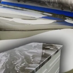 Wylewanie ciemnej żywicy na jasną powierzchnię, tworząc efekt marmuru na blacie szafki. Błękitna taśma malarska w tle. Detal procesu aplikacji.