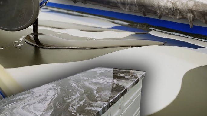 Wylewanie ciemnej żywicy na jasną powierzchnię, tworząc efekt marmuru na blacie szafki. Błękitna taśma malarska w tle. Detal procesu aplikacji.