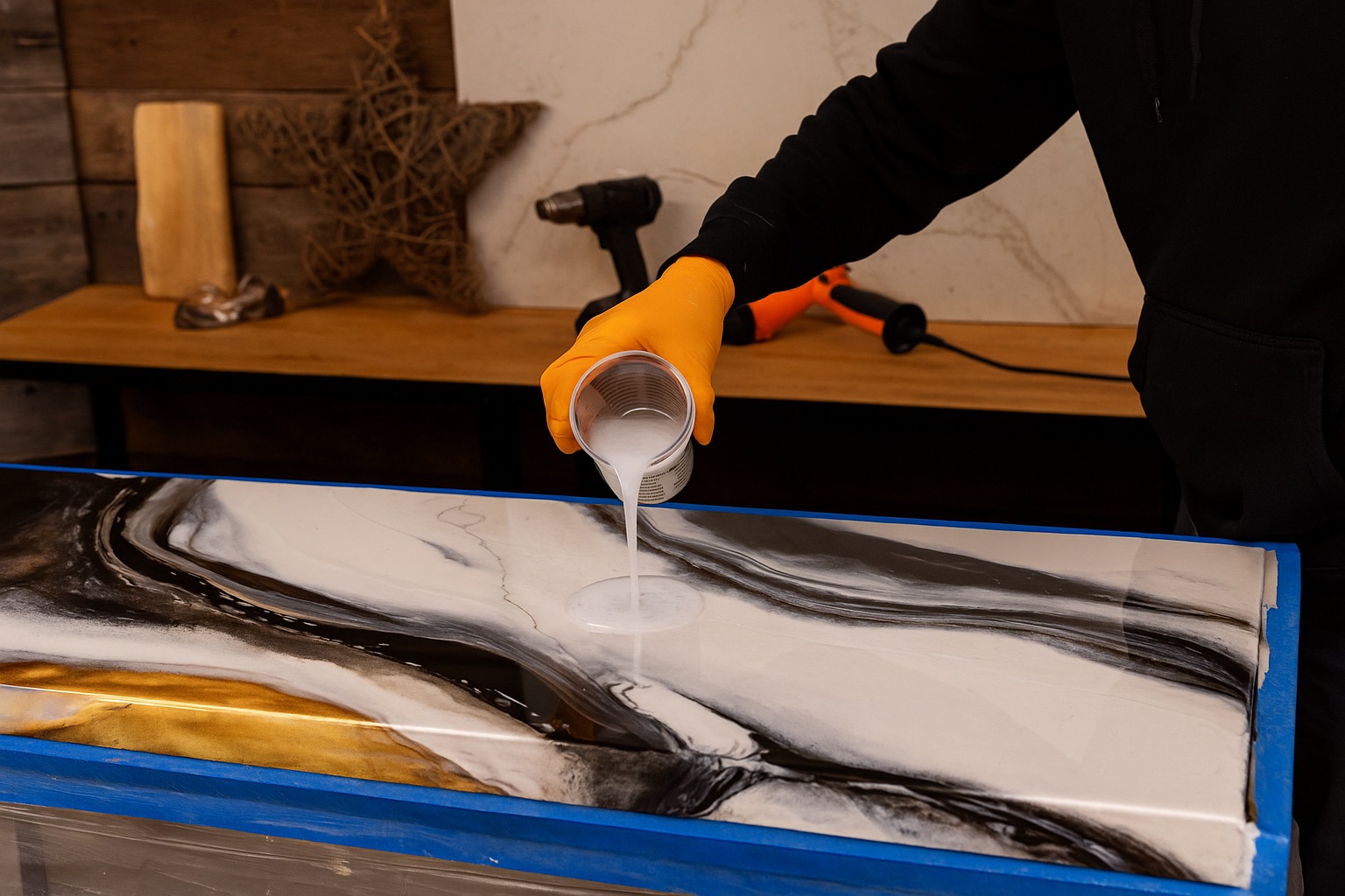 Wylewanie żywicy na blat z efektem marmuru w odcieniach bieli, czerni i złota. Blat zabezpieczony niebieską taśmą. W tle narzędzia i dekoracje.