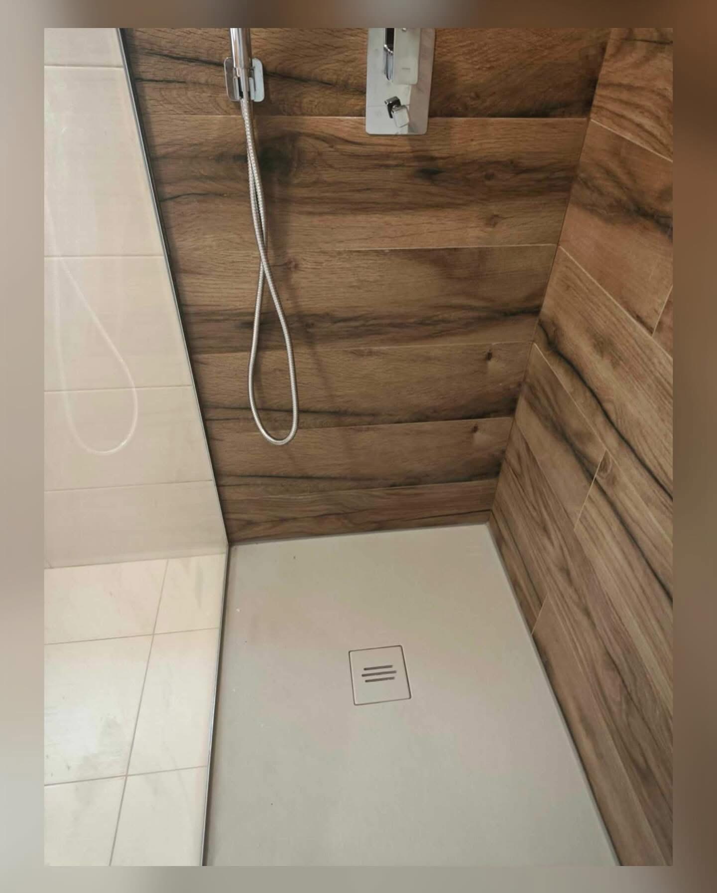 Nowoczesna kabina prysznicowa z brązowymi płytkami imitującymi drewno na ścianach i białą, kwadratową brodzikiem z odpływem liniowym.