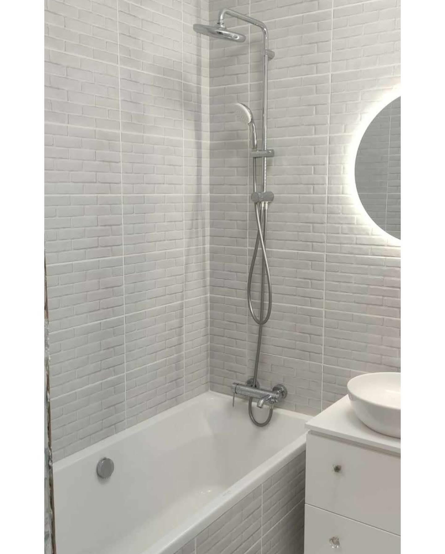 Wykończona łazienka z wanną i umywalką na szafce. Ściany wyłożone szarą, cegiełkową glazurą. Nad wanną zestaw prysznicowy. Okrągłe lustro z podświetleniem.