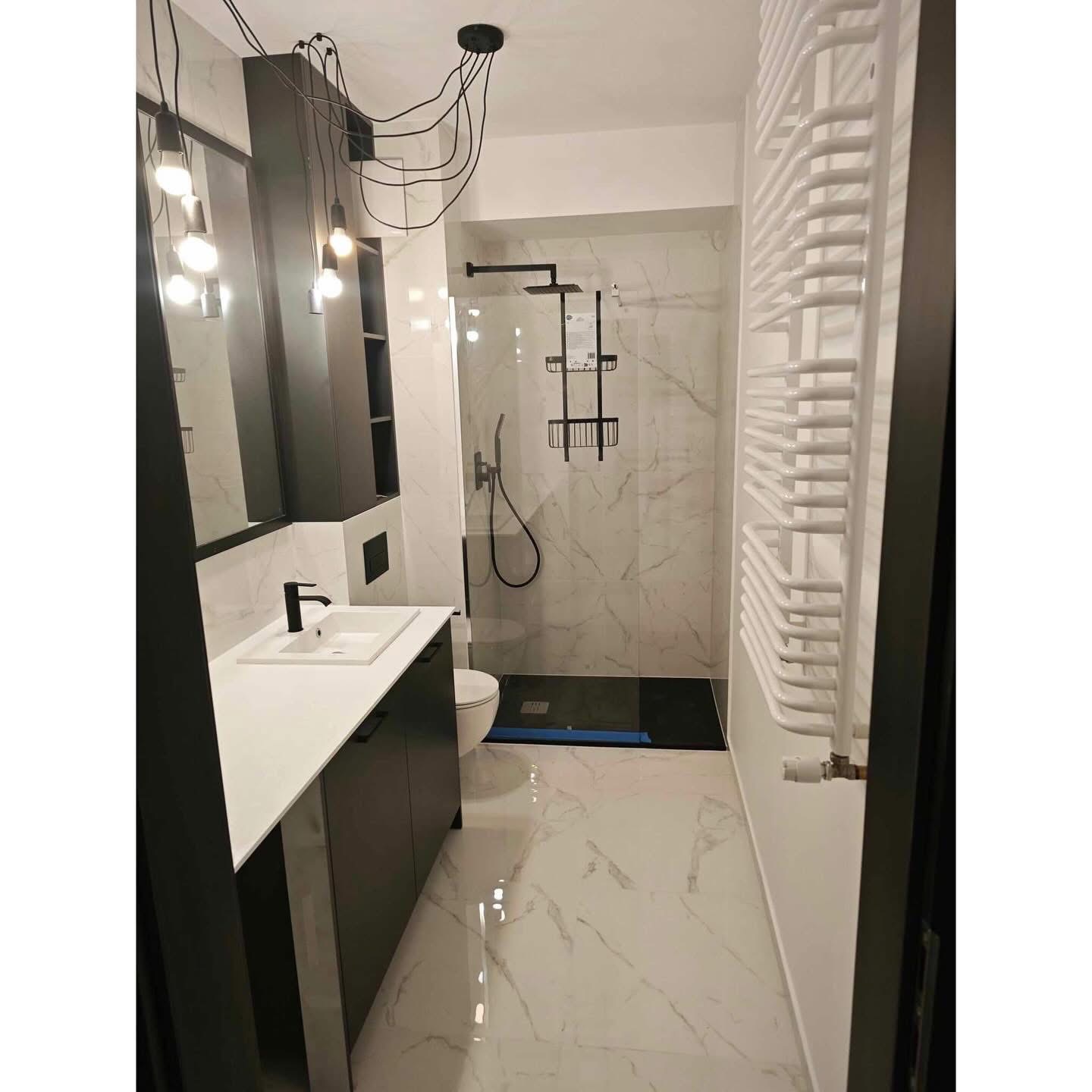Nowoczesna łazienka w bieli i czerni: marmurowe płytki, kabina prysznicowa typu walk-in, wisząca szafka z umywalką i dekoracyjne oświetlenie sufitowe.