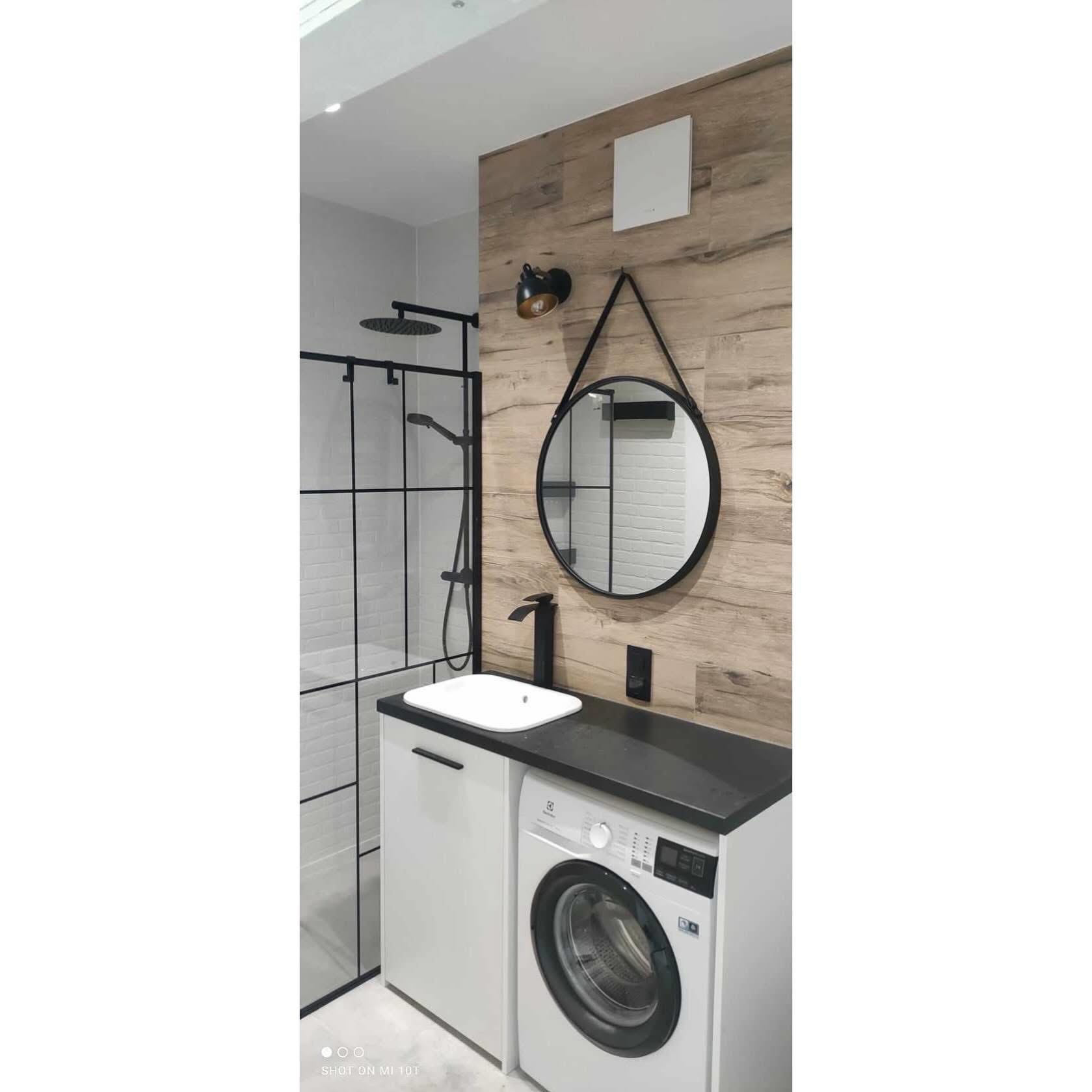 Nowoczesna łazienka z kabiną typu walk-in, umywalką nablatową, pralką i okrągłym lustrem na tle ściany z płytek imitujących drewno. Minimalistyczny design w odcieniach bieli, czerni i drewna.