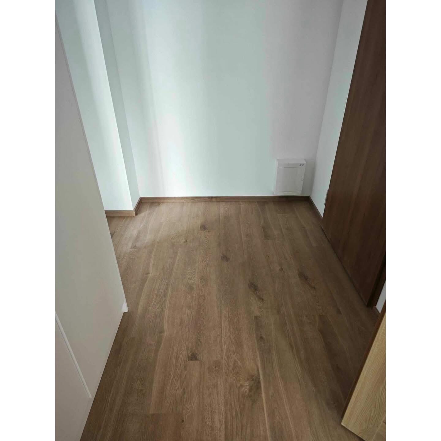 Jasny pokój z drewnianą podłogą ułożoną w jodełkę, białe ściany i brązowe drzwi. Widoczny fragment szafki elektrycznej na ścianie.