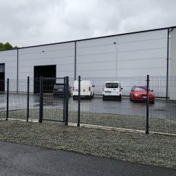 VessTech Group Sp. z o.o. - Nowoczesna hala produkcyjna z szarej blachy, ogrodzona metalową siatką. Na parkingu zaparkowane samochody. Tabliczka z adresem firmy.