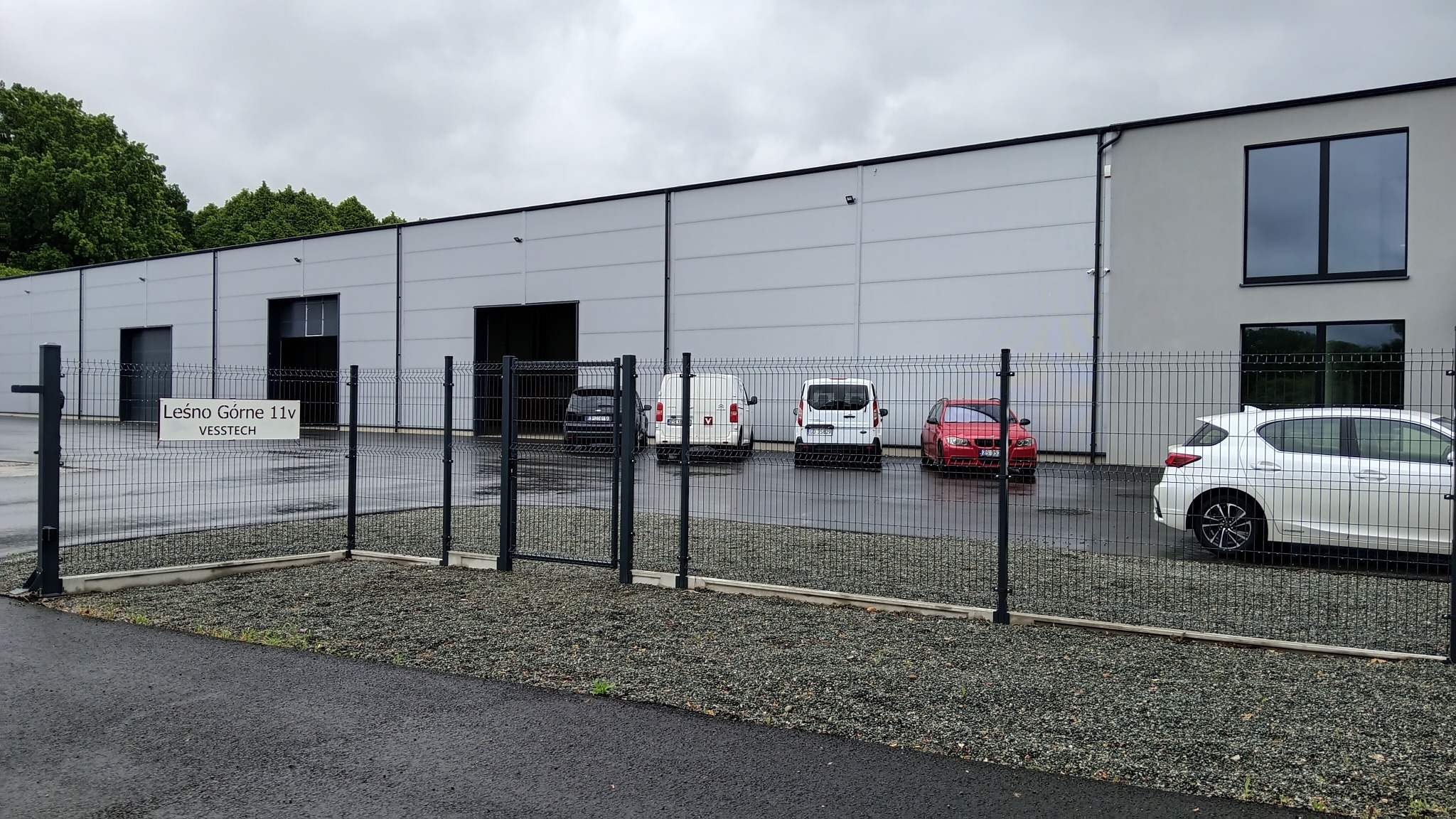 Nowoczesna hala produkcyjna z szarej blachy, ogrodzona metalową siatką. Na parkingu zaparkowane samochody. Tabliczka z adresem firmy.